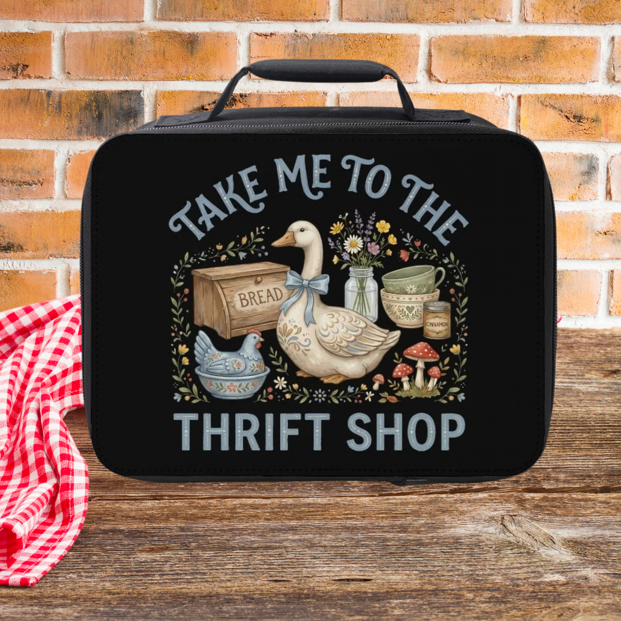 Black lunch bag with a goose design and text 'Take Me to the Thrift Shop' on a wooden surface with a brick wall background.
