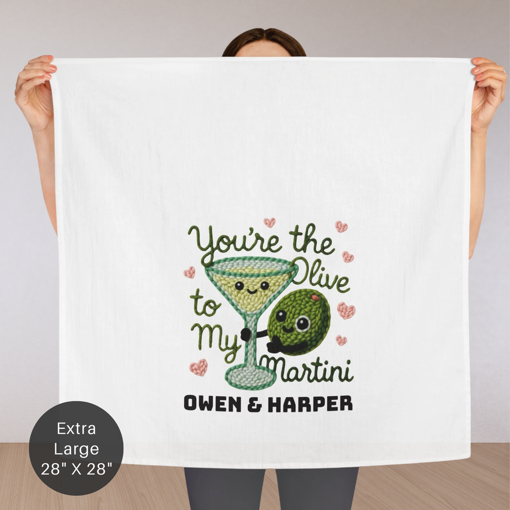 Person holding a decorative towel with a cartoon olive and martini glass design, text 'You're the Olive to my Martini' and names 'Owen & Harper'.