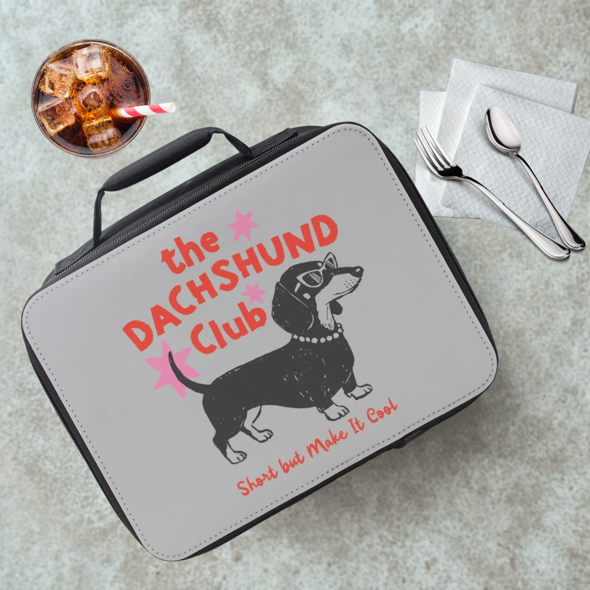 Lunch box with dachshund design and 'Dachshund Club' text on a cafeteria table with a drink and utensils