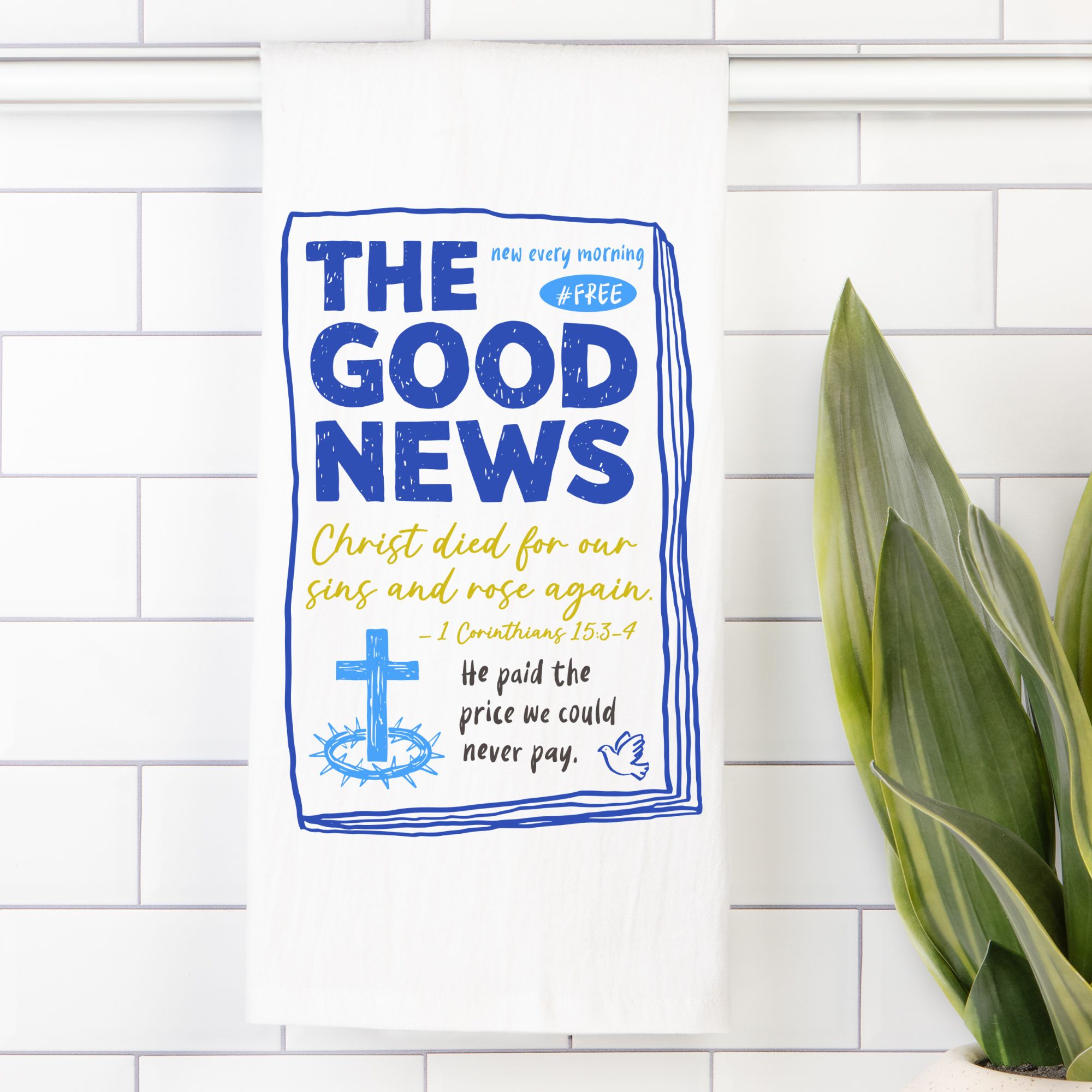 Tea towel with 'The Good News' text on a white tiled wall with a plant.