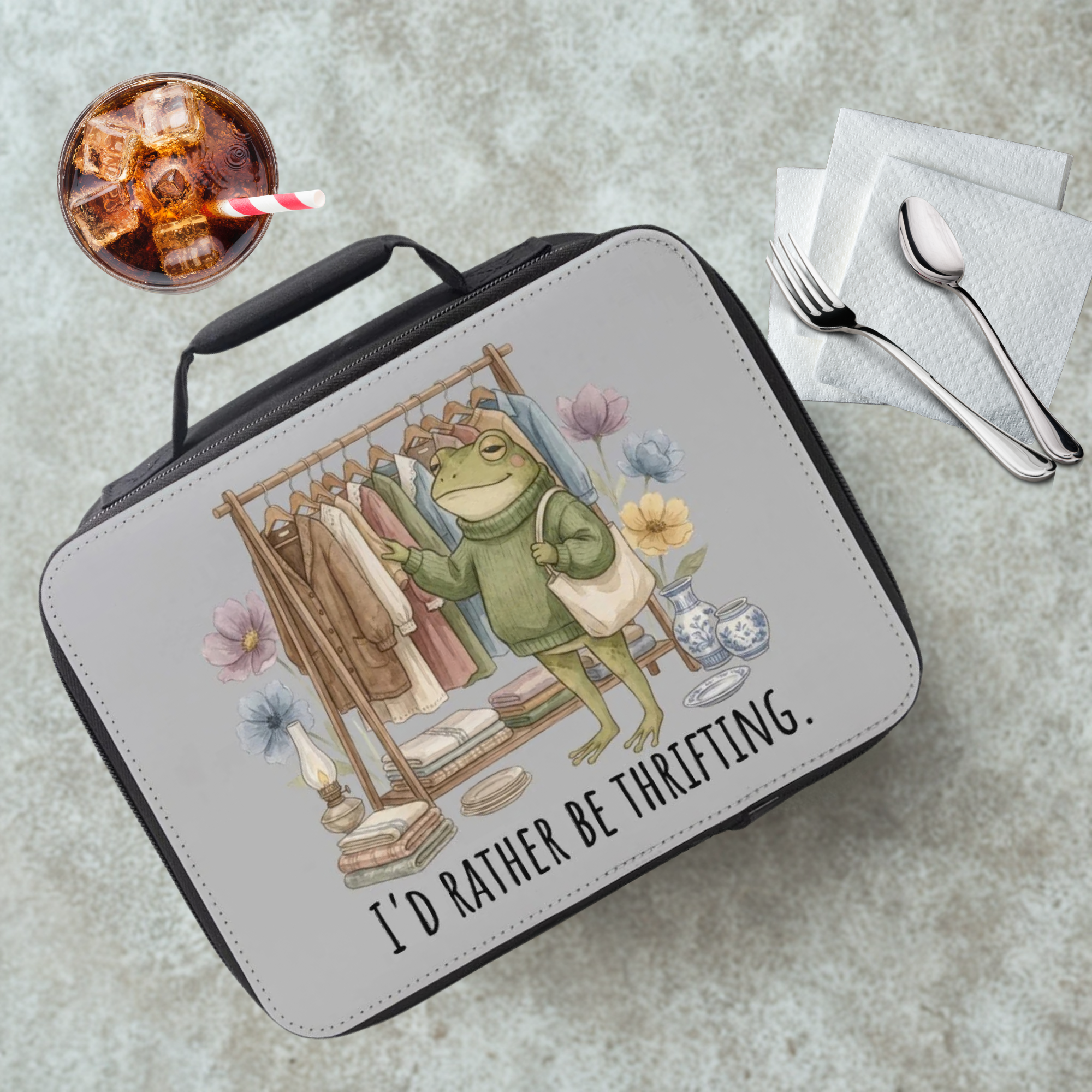 Lunch bag with a frog graphic and text 'I'd rather be thrifting.' on a cafeteria table with a drink and utensils.