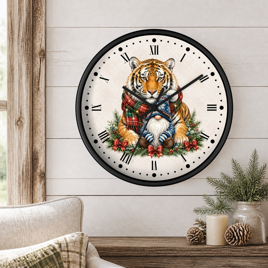 A round wall clock with a black frame and a white face featuring an illustrated tiger wearing a red plaid scarf, seated among greenery and red berries. The clock is shown hanging on a light wood plank wall above a wooden table with a candle, pinecones, and greenery, with part of a beige chair visible in the lower left.