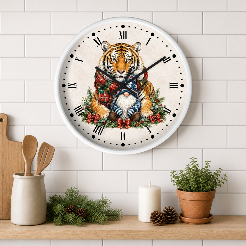 A round wall clock with a white face and Roman numerals, featuring an illustration of a tiger wrapped in a red tartan plaid scarf , sitting among green pine branches and red berries. The clock is mounted on a white subway tile wall above a wooden shelf styled with kitchen utensils, candles, pinecones, and a potted plant.