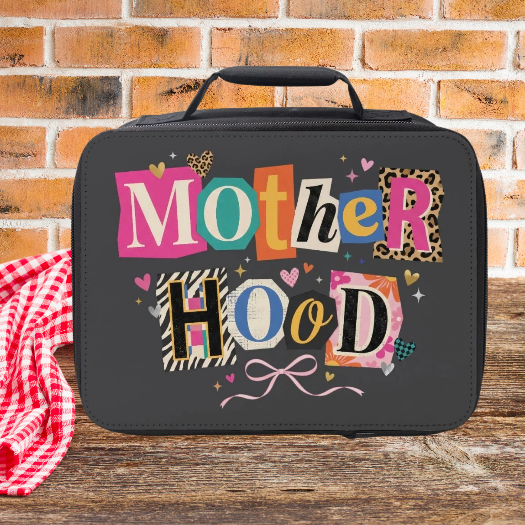 Black insulated lunch bag with colorful 'Mother Hood' text on a wooden picnic table against a brick wall background