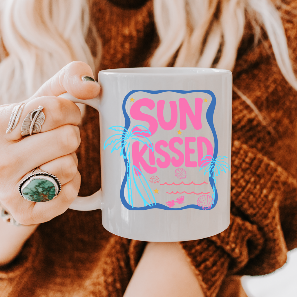 Person holding an 11oz. mug with 'Sun Kissed' design, wearing a brown sweater and rings.