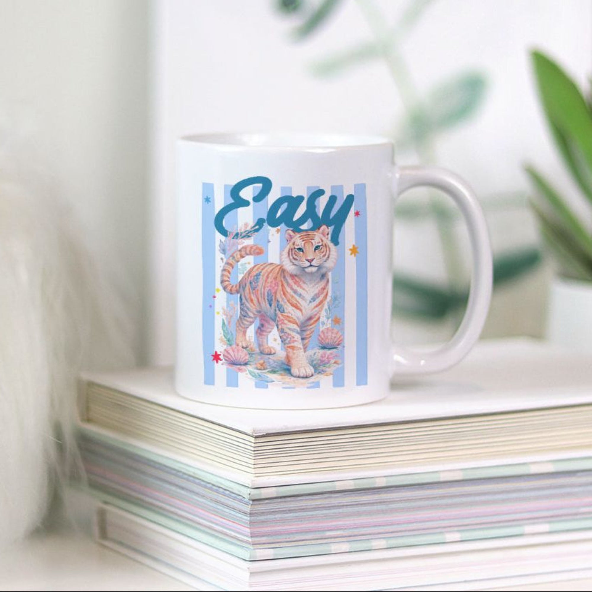 White 11oz mug with a tiger design and 'Easy' text on a stack of books with plants in the background