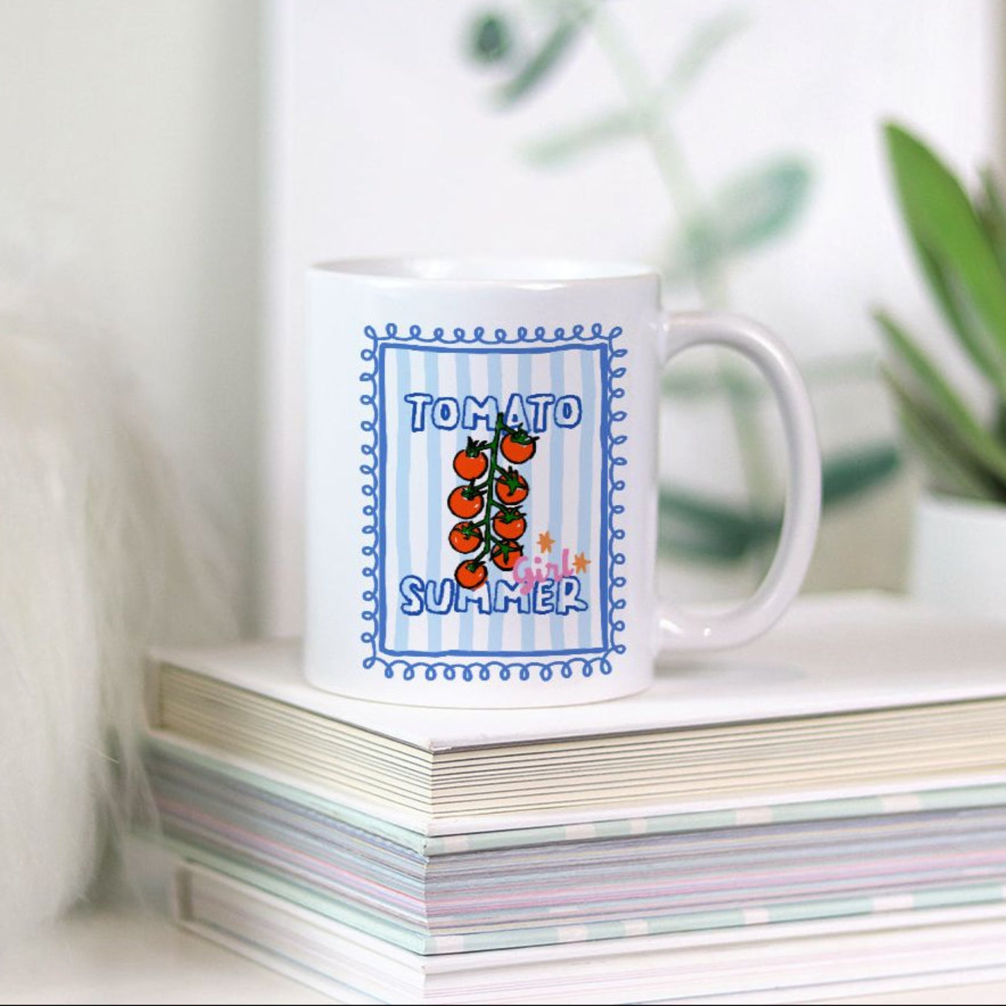 White 11oz. mug with tomato design and 'Tomato Summer' text on a stack of books with plants in the background