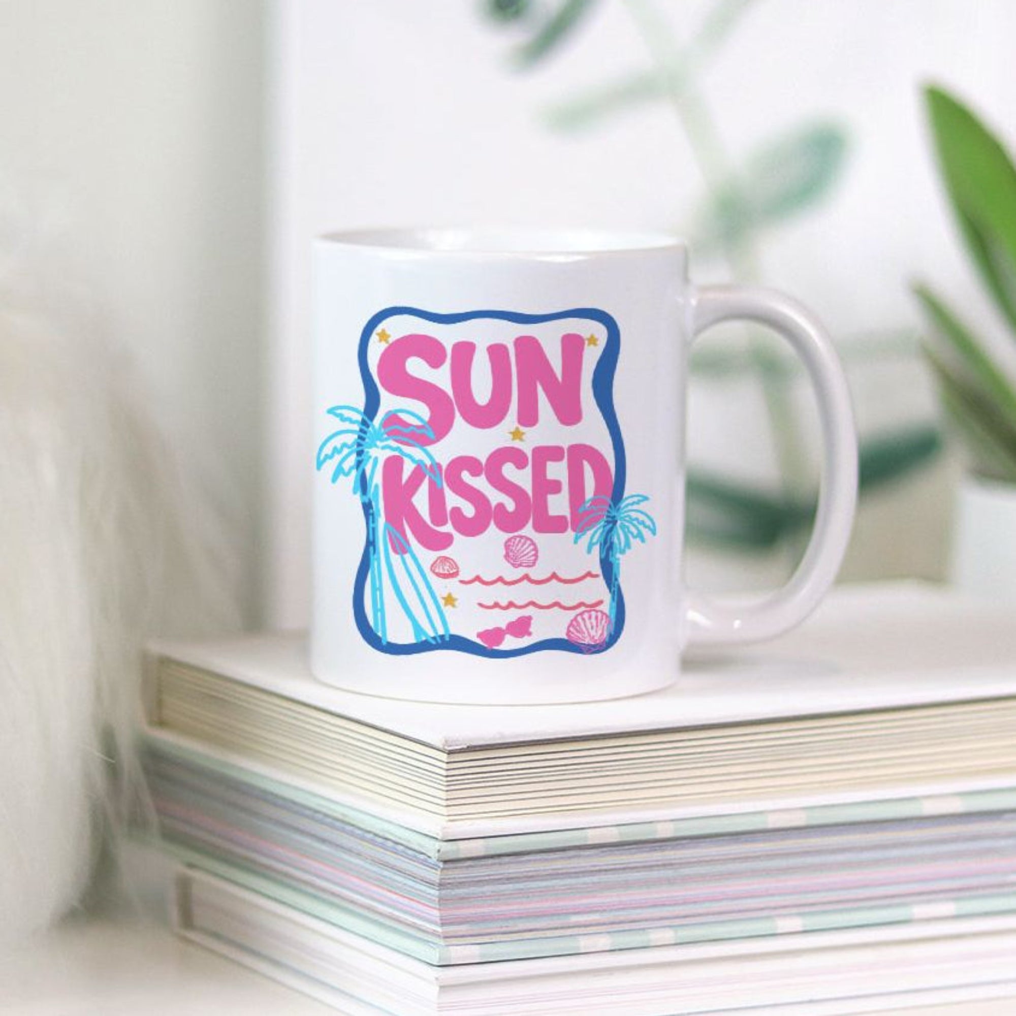 White 11oz mug with 'Sun Kissed' design on a stack of books with a plant in the background