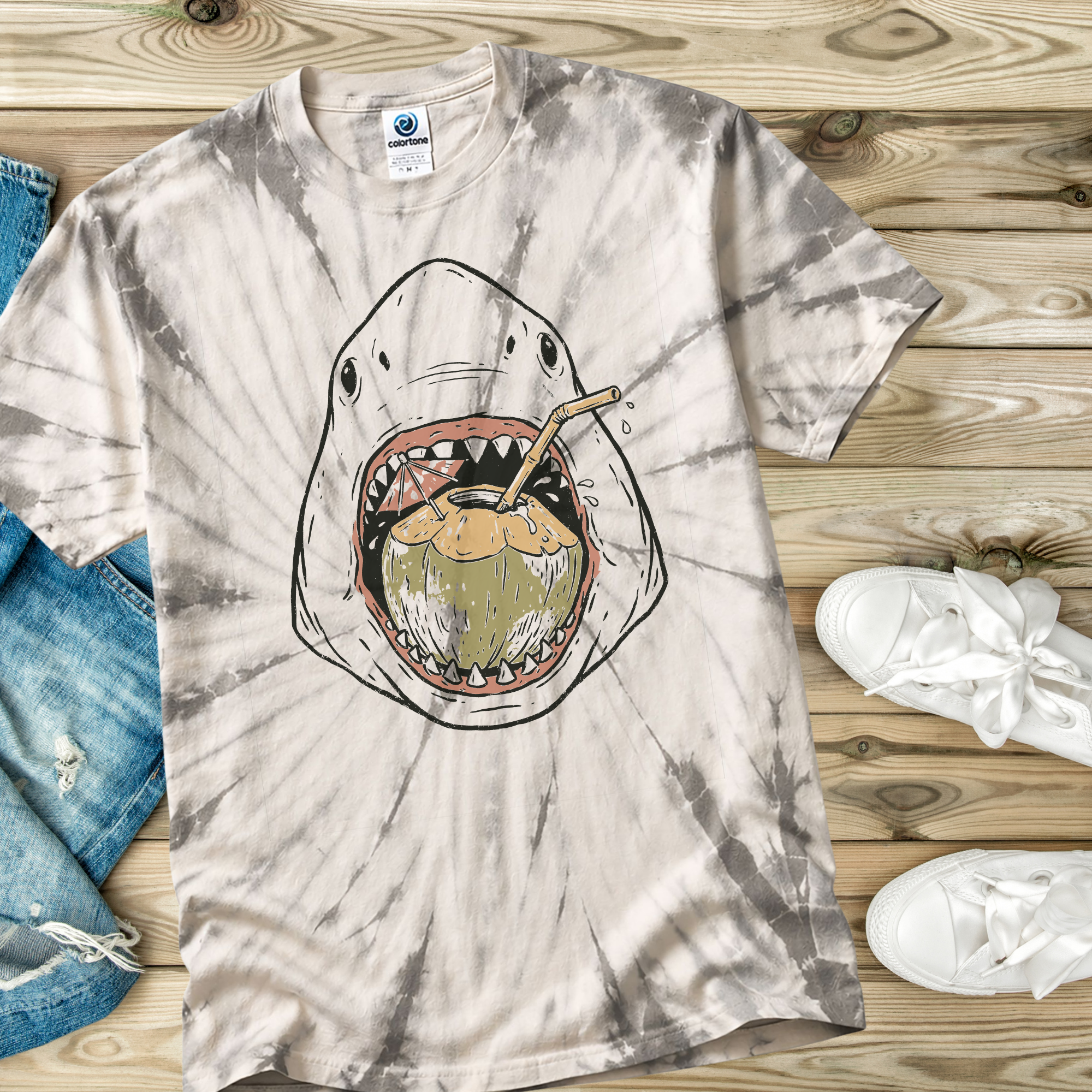 Tie-dye t-shirt in the color Spider Silver with a shark graphic on a wooden surface with jeans and sneakers.