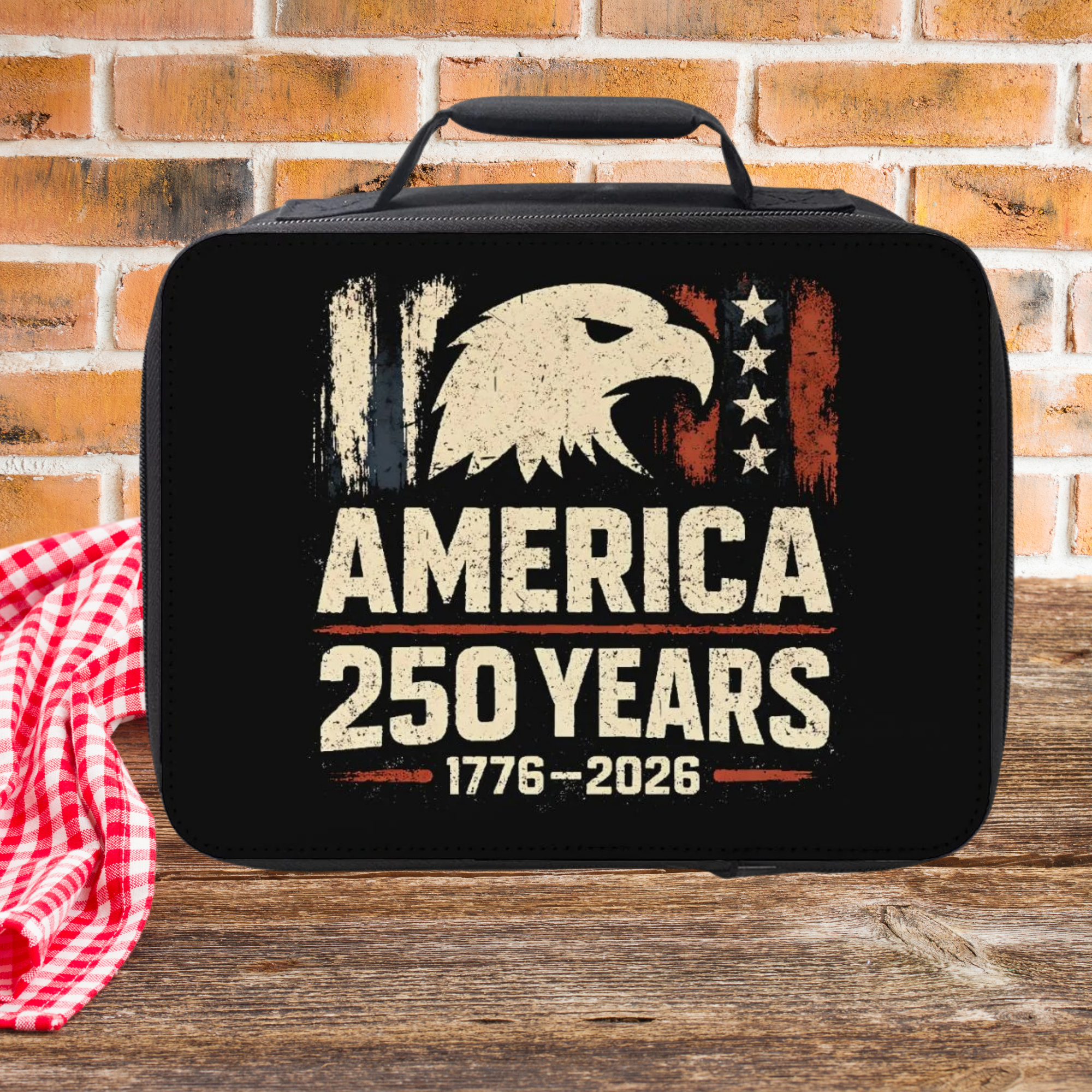 Black lunch bag with American eagle design and 'America 250 Years' text on a picnic table against a brick wall background