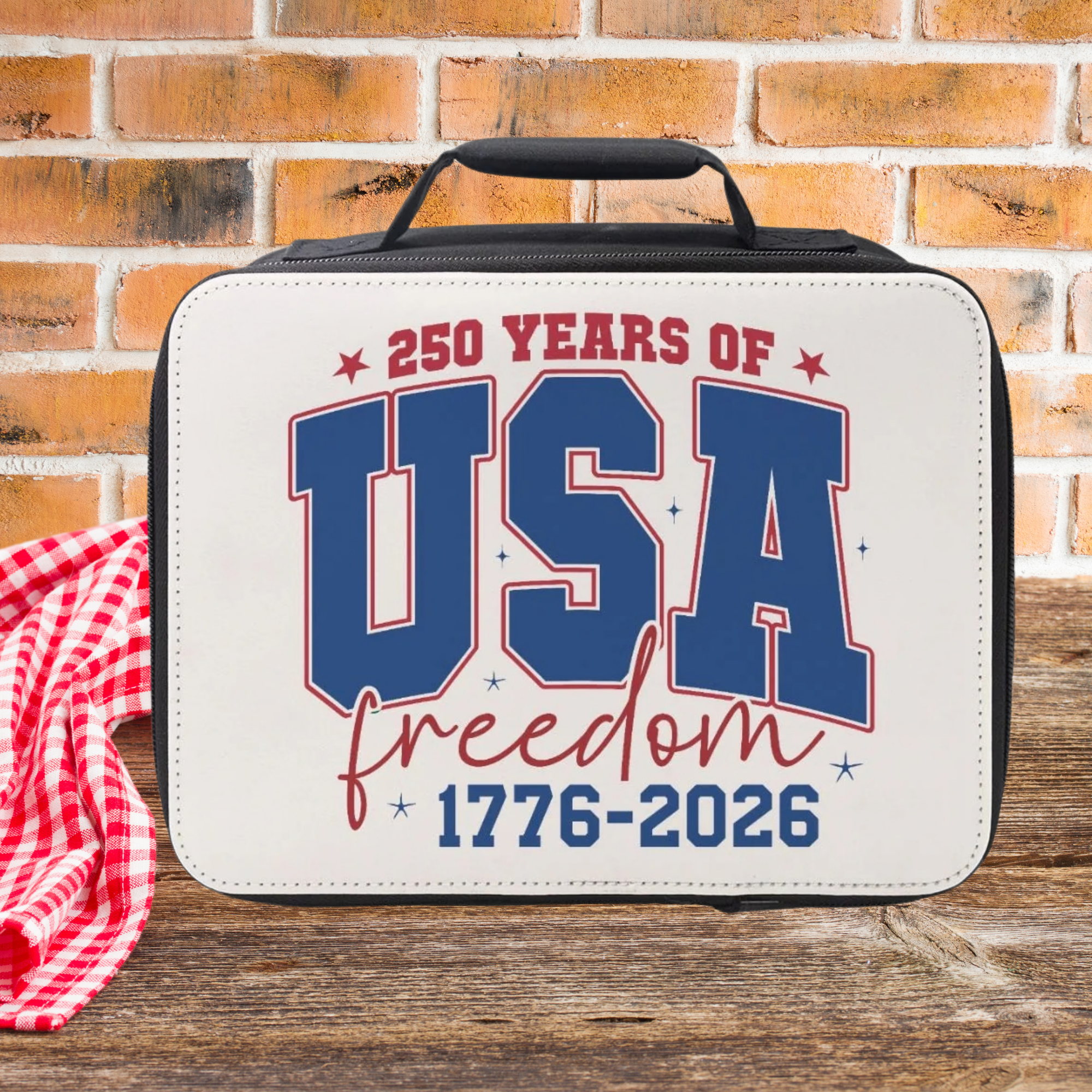 insulated lunch bag with '250 Years of USA Freedom 1776-2026' text on a wooden picnic table with a brick wall background.