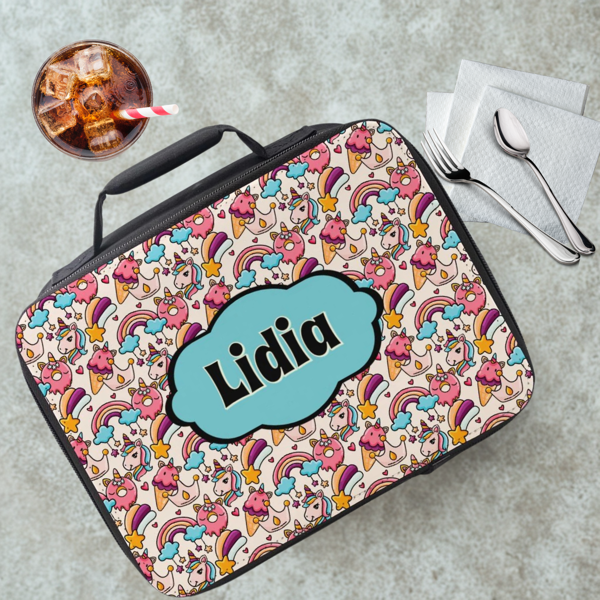 Colorful unicorn patterned lunch bag with 'Lidia' custom name on a gray table with a drink and utensils.