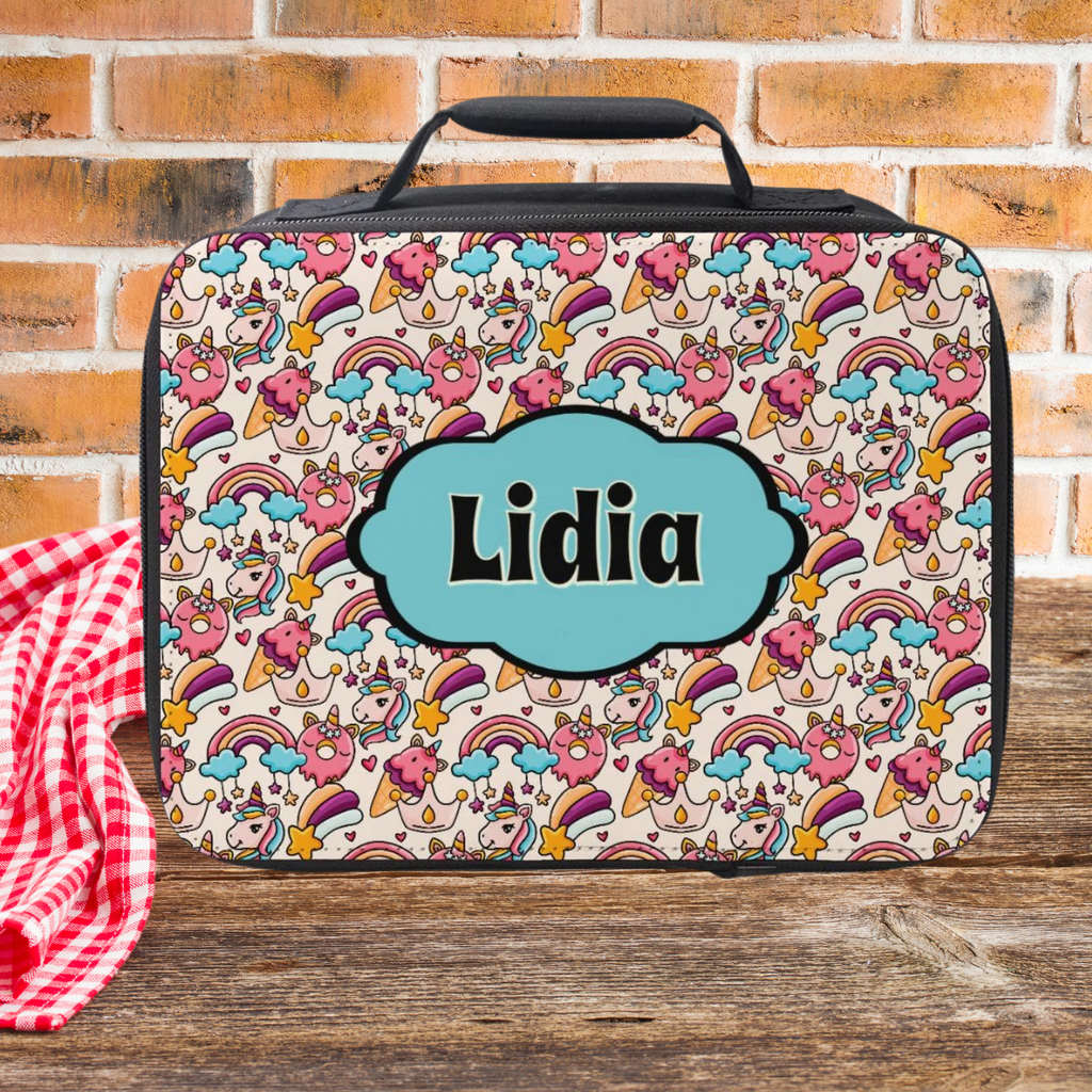 Colorful unicorn rainbow patterned lunch bag with 'Lidia' custom text on a wooden picnic table against a brick wall background