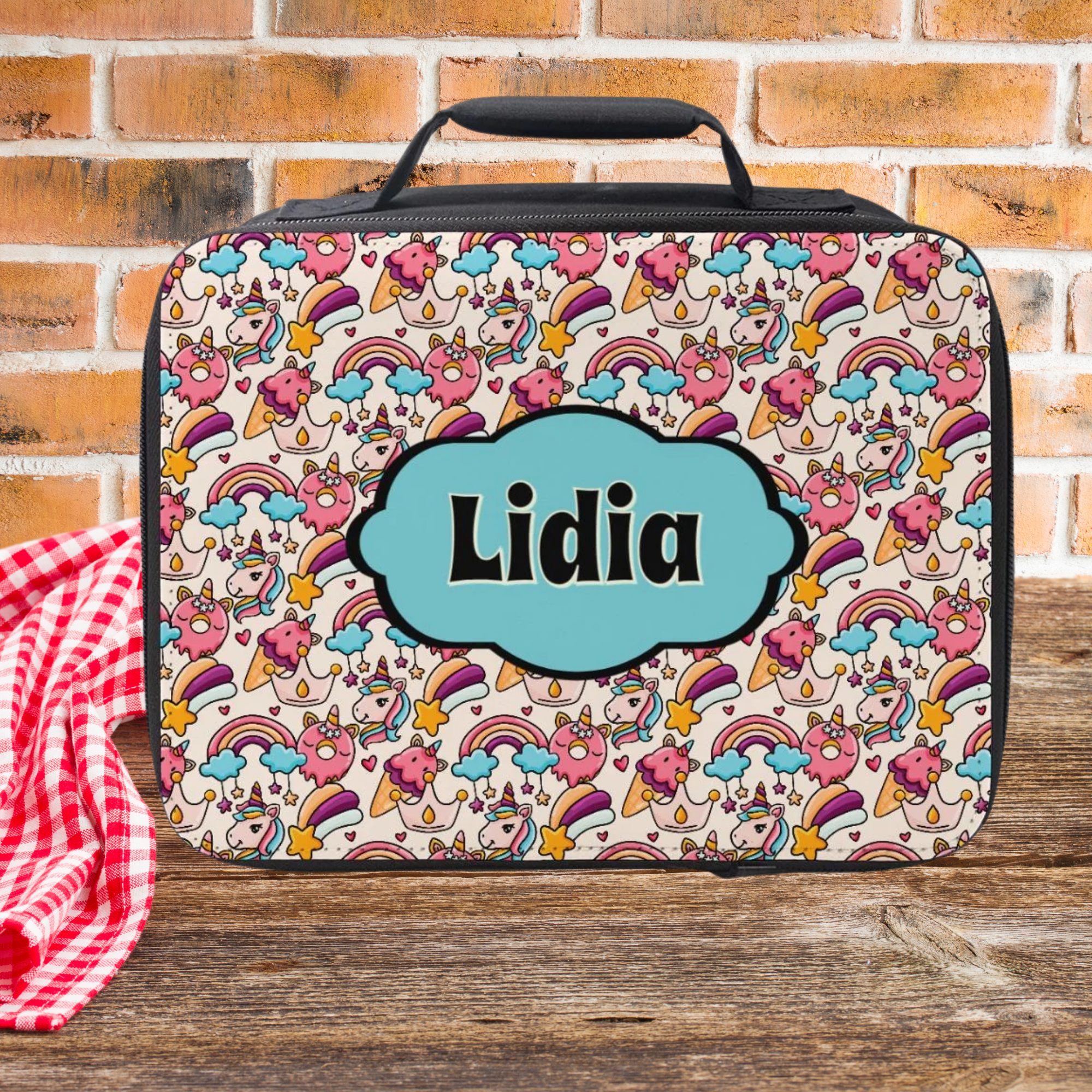 Colorful unicorn rainbow patterned lunch bag with 'Lidia' custom text on a wooden picnic table against a brick wall background