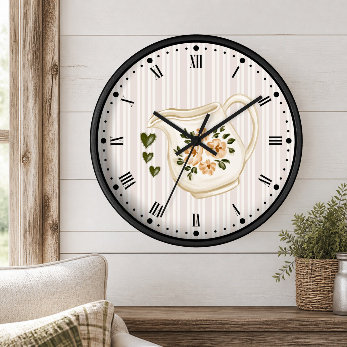 A round wall clock with a black frame and a white face featuring a cream-colored pitcher illustration, green hearts, and floral details. The clock is mounted on a white shiplap wall above a wooden shelf with a potted plant and part of a sofa visible.