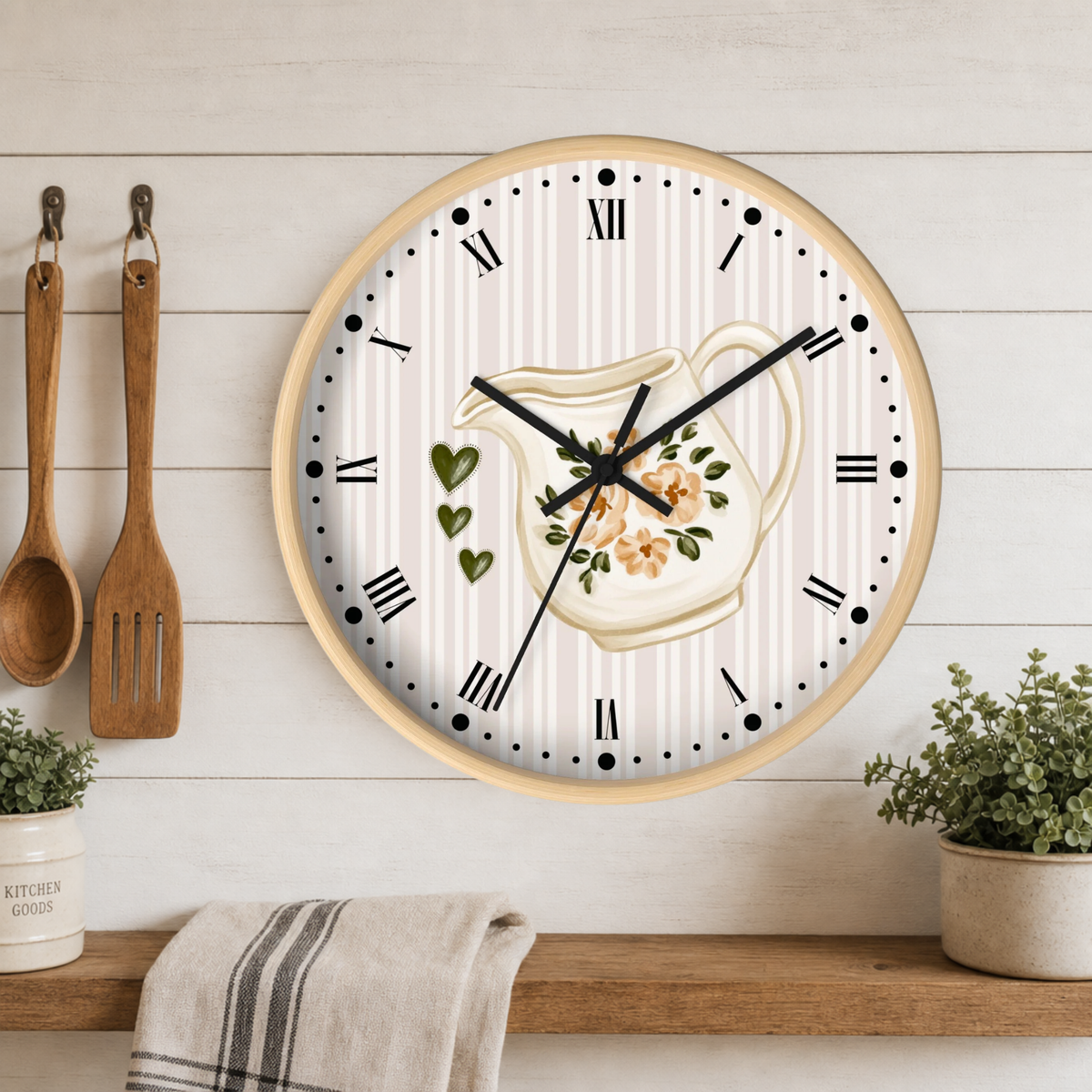 A round wall clock with a light wood frame hangs on a white wood-paneled wall above a wooden shelf. The clock face has a cream and white striped background with a central illustration of a floral pitcher, black Roman numerals, and black clock hands.