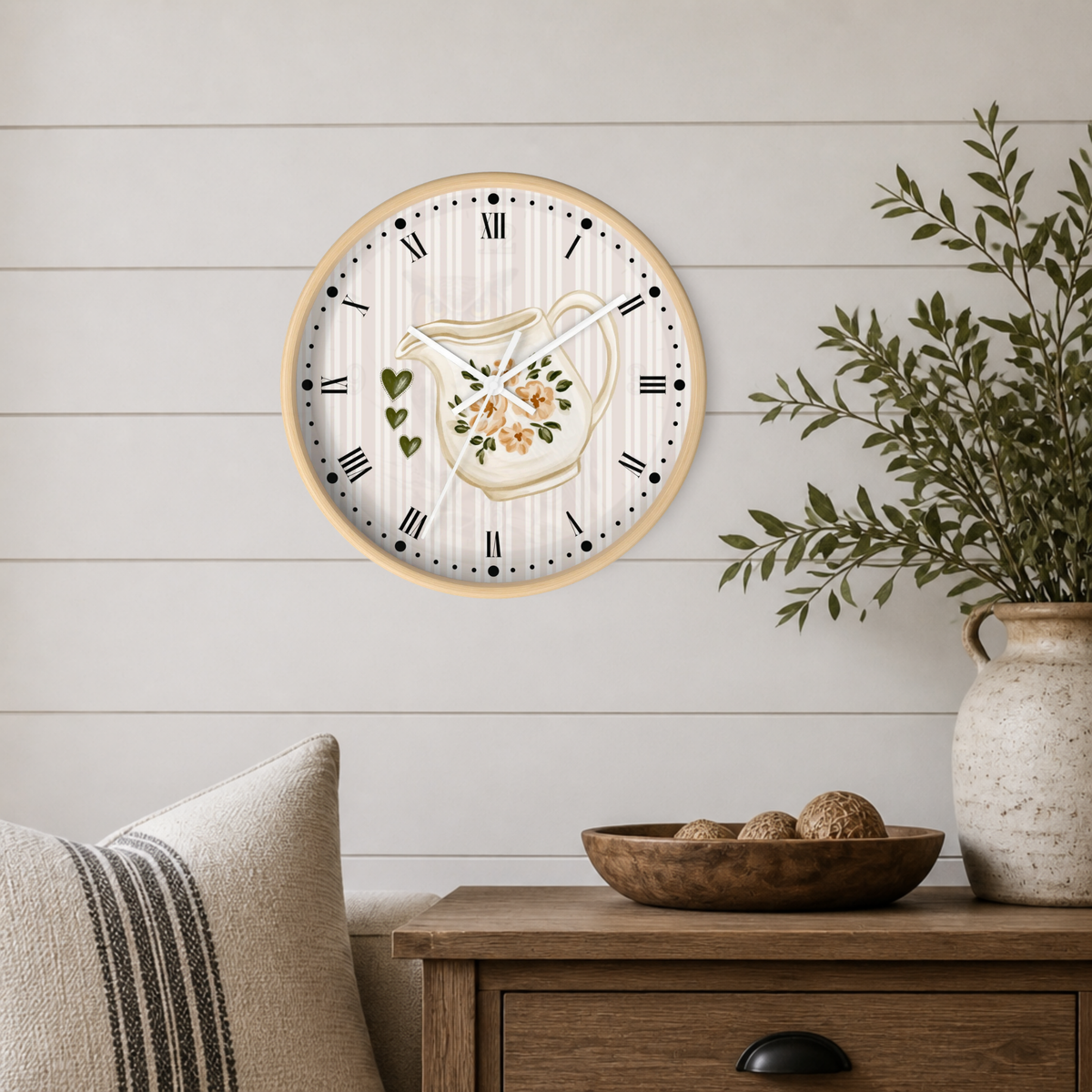 A round wall clock with a light wood frame hangs on a white shiplap wall above a wooden side table. The clock face is white with vertical stripes, black Roman numerals, and a central floral pitcher illustration.