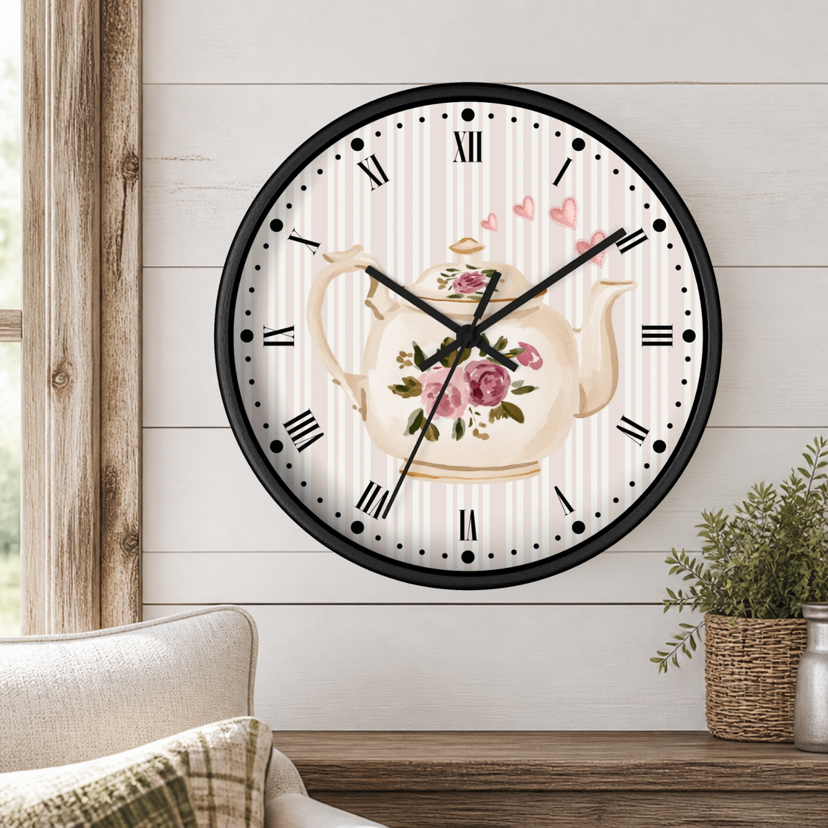 A round wall clock with a black frame and a floral teapot design on the face, mounted on a white shiplap wall above a wooden mantel with a small potted plant.
