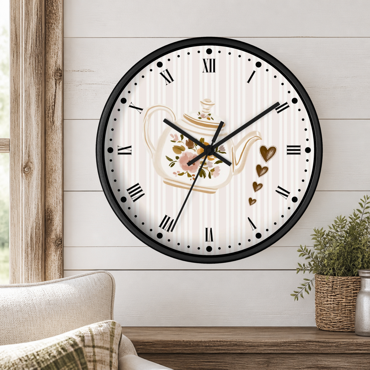 A round wall clock with a black frame and a white face featuring a floral teapot illustration, Roman numerals, and gold heart accents, mounted on a white shiplap wall above a wooden table with a plant.