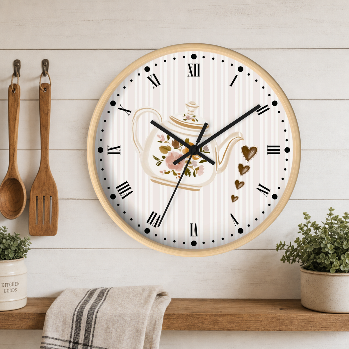 Round wall clock with a light wood frame and a white face featuring a floral teapot illustration, Roman numerals, and black hands, displayed on a wooden shelf against a white paneled wall with kitchen utensils and small plants.