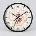 Round wall clock with a black frame and a tea-themed face featuring a cream teapot with pink roses, Roman numerals, and a light striped background.