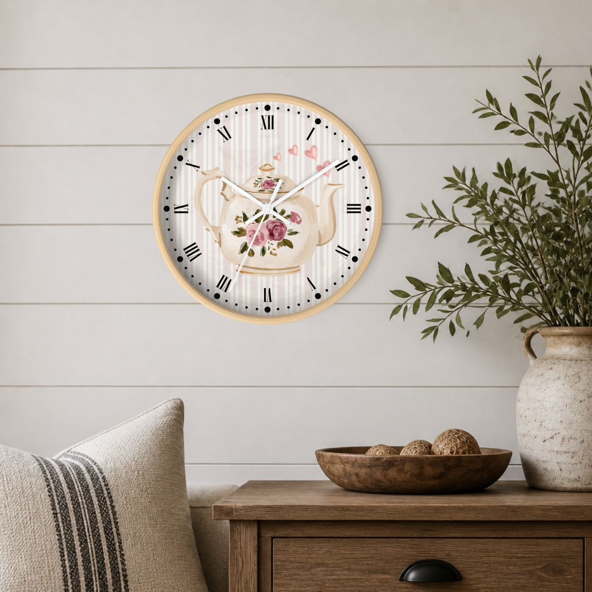 Round wall clock with a light wood frame and a floral teapot illustration on the face, mounted on a white shiplap wall above a wooden side table with a vase and pillows.