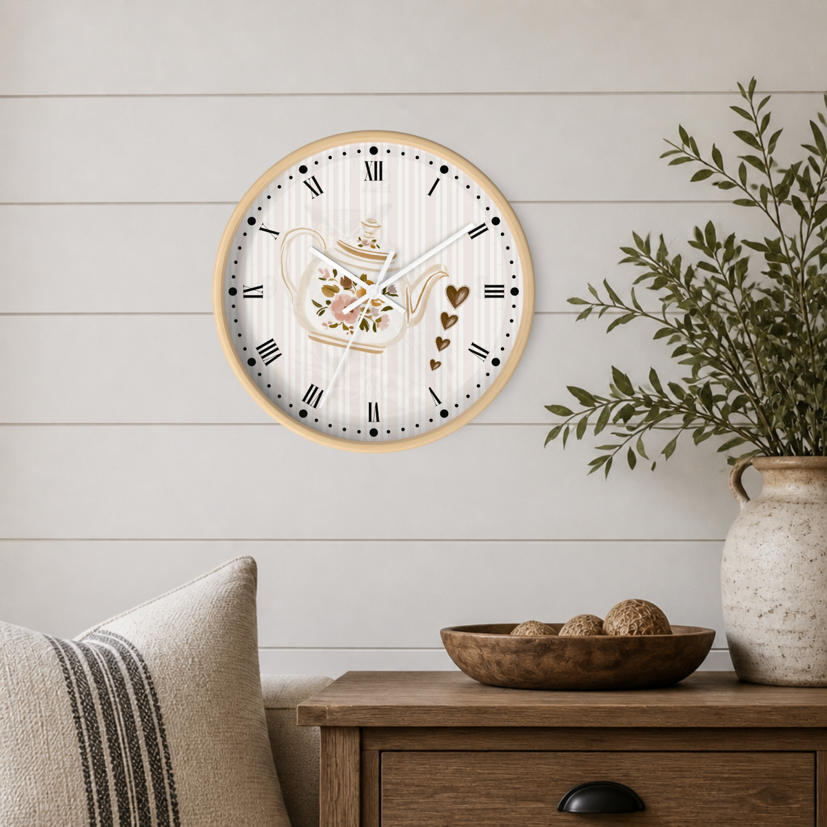 A round wall clock with a light wood frame and a white face featuring a floral teapot illustration and black Roman numerals, styled on a white shiplap wall above a wooden side table with a plant and decorative bowl.