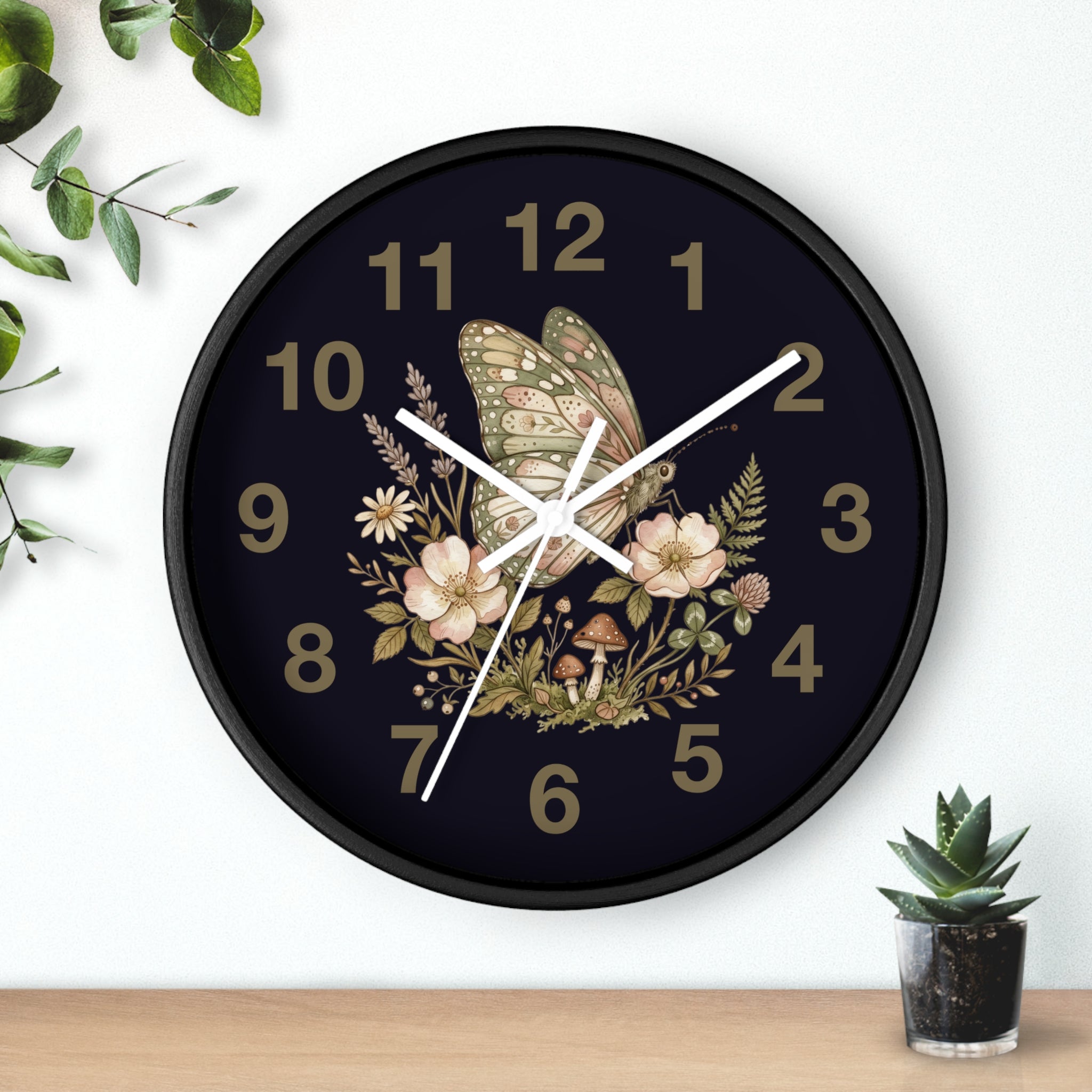 Decorative round black wall clock with white hands and butterfly and floral design on a dark background, placed on a wooden surface with a plant.