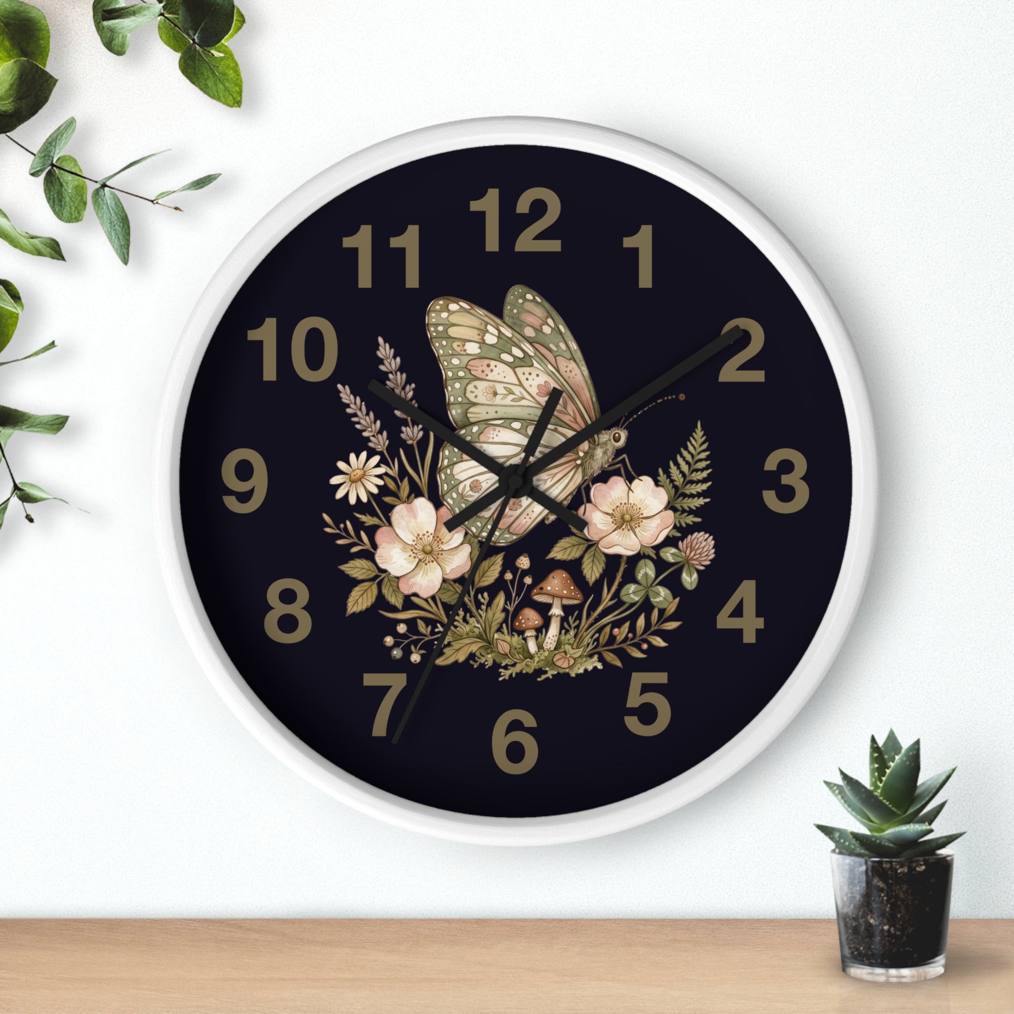 Decorative round white wall clock with black hands and butterfly and floral design on a black background, placed on a white surface with a plant and small pot in the foreground.