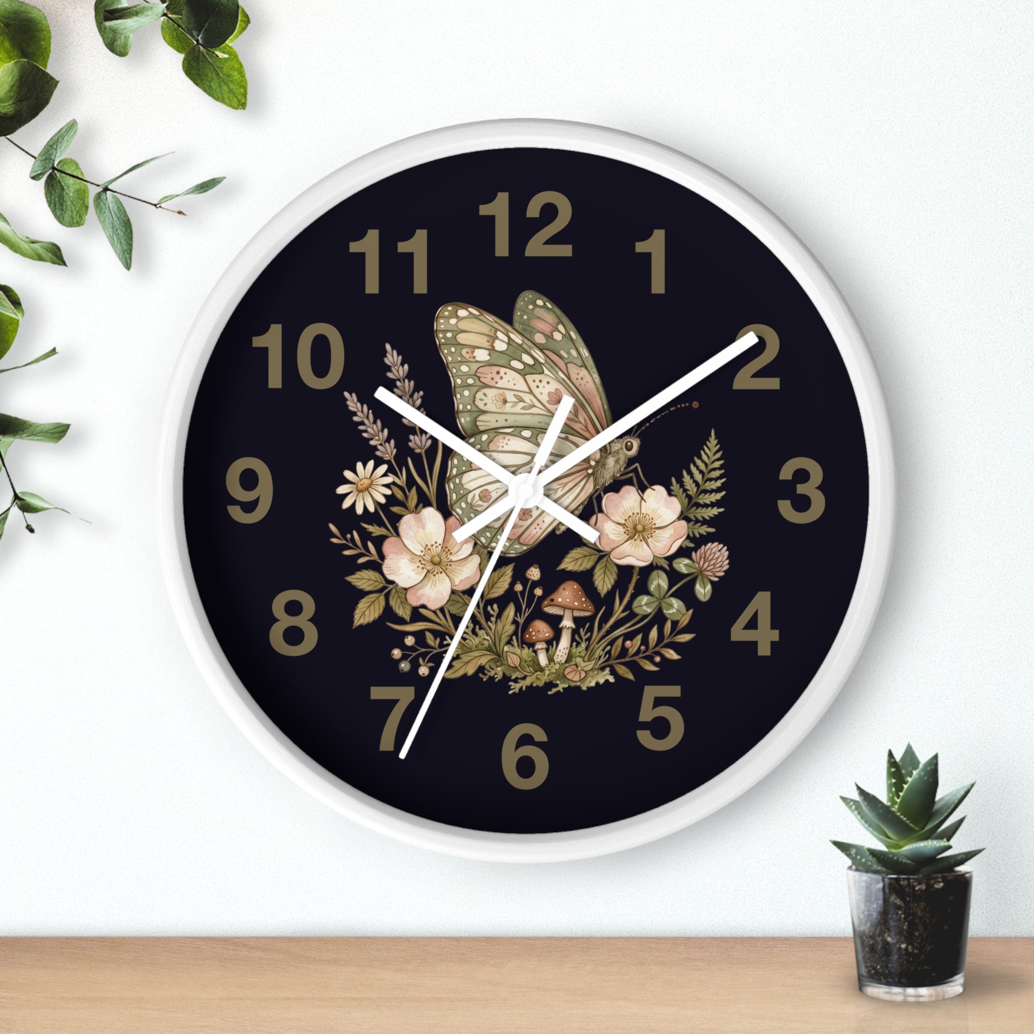 Decorative round white wall clock with butterfly and floral design on a dark background, placed on a wooden surface with a plant.