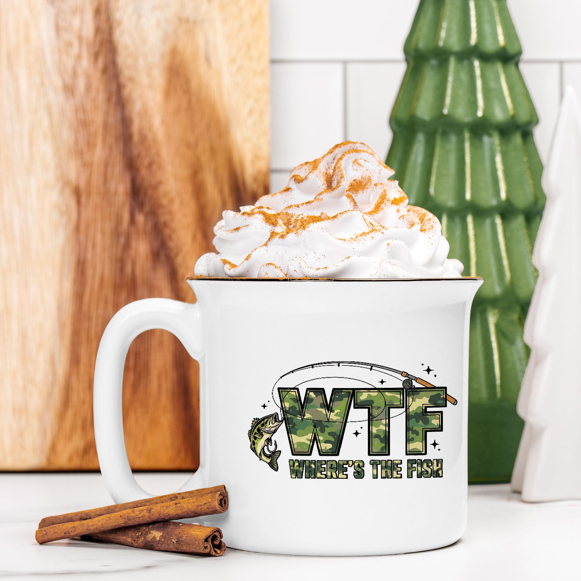 White mug with 'WTF Where's the Fish' design, topped with whipped cream and a cinnamon stick, against a wooden and green decorative background.