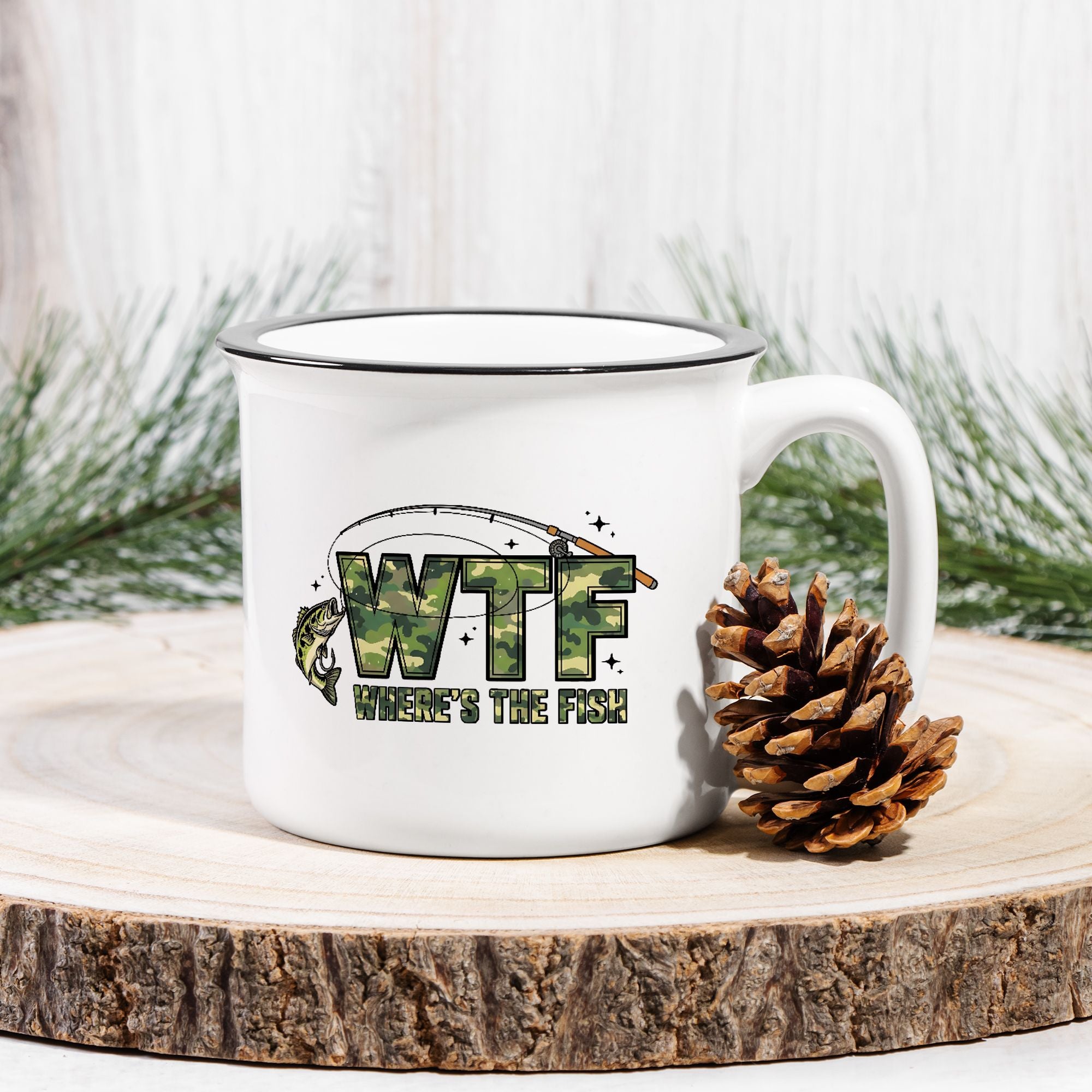 White mug with 'WTF Where's the Fish?' design on a wooden surface with a pine cone and evergreen branches.