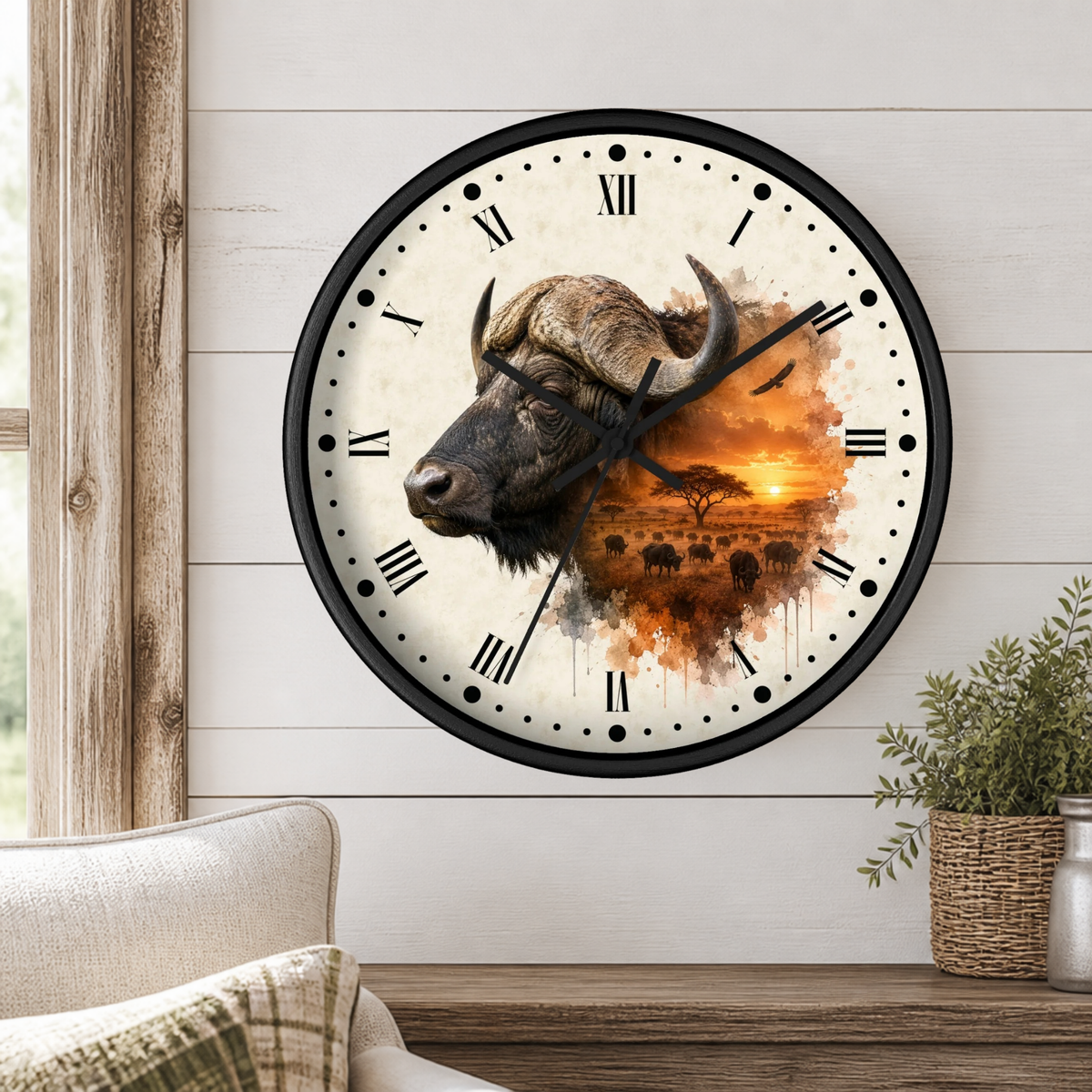A round wall clock with a black frame and Roman numerals, featuring a watercolor-style print of an African buffalo head over an orange savanna sunset. The clock is mounted on a white wood-paneled wall above a wooden console table with a small potted plant and part of a sofa visible.