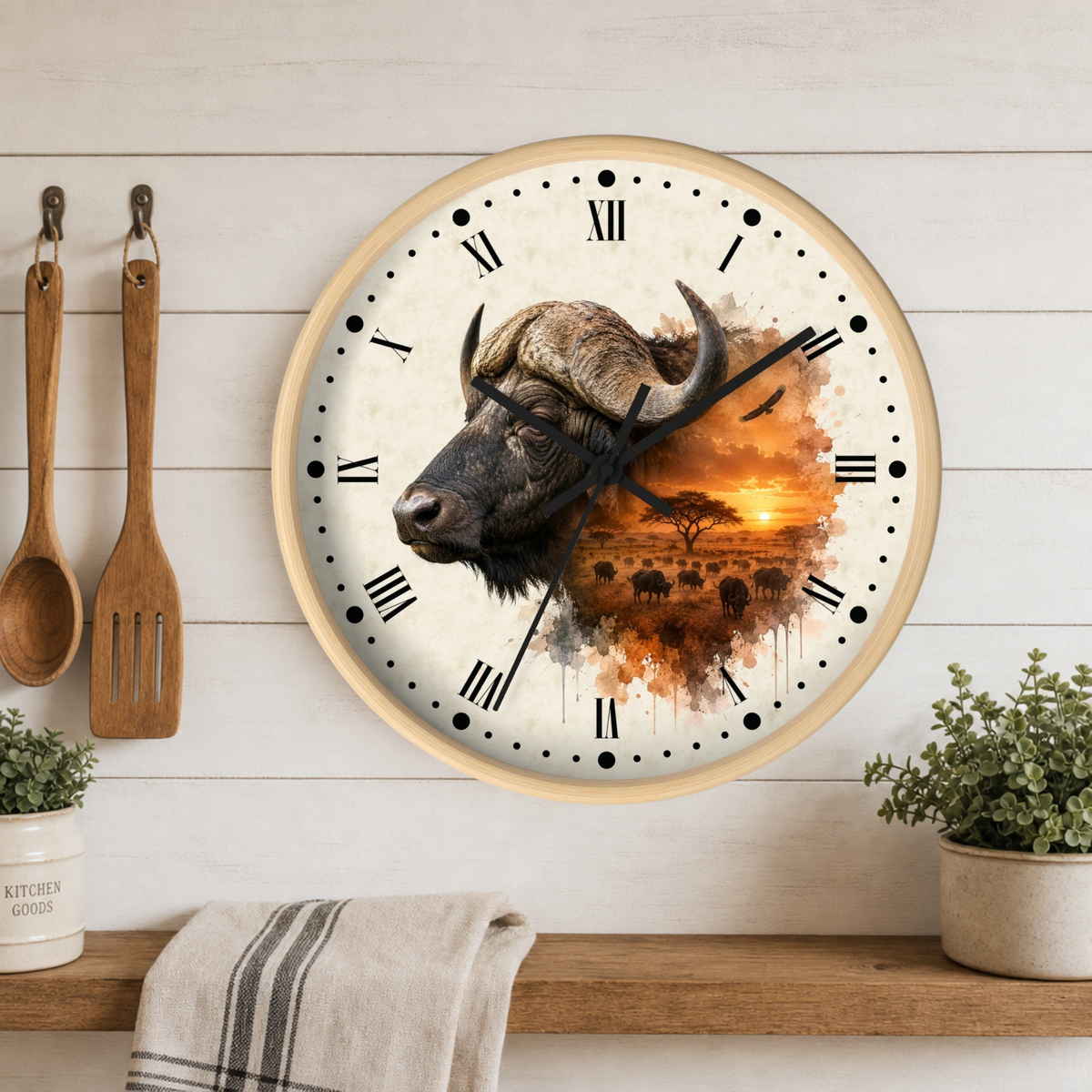 A round wall clock with a light wood frame hangs on a white wood-paneled wall above a shelf. The clock face features a watercolor-style illustration of a buffalo head on the left and an orange sunset landscape with acacia trees on the right.