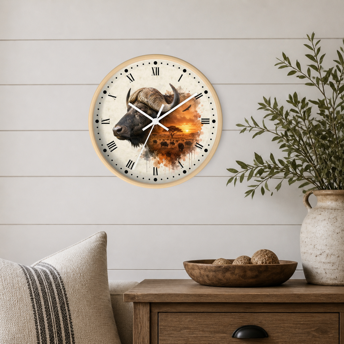 A round wall clock with a light wood frame hangs on a white shiplap wall above a wooden side table. The clock face features a detailed illustration of an African buffalo in profile with a warm orange sunset landscape behind it.