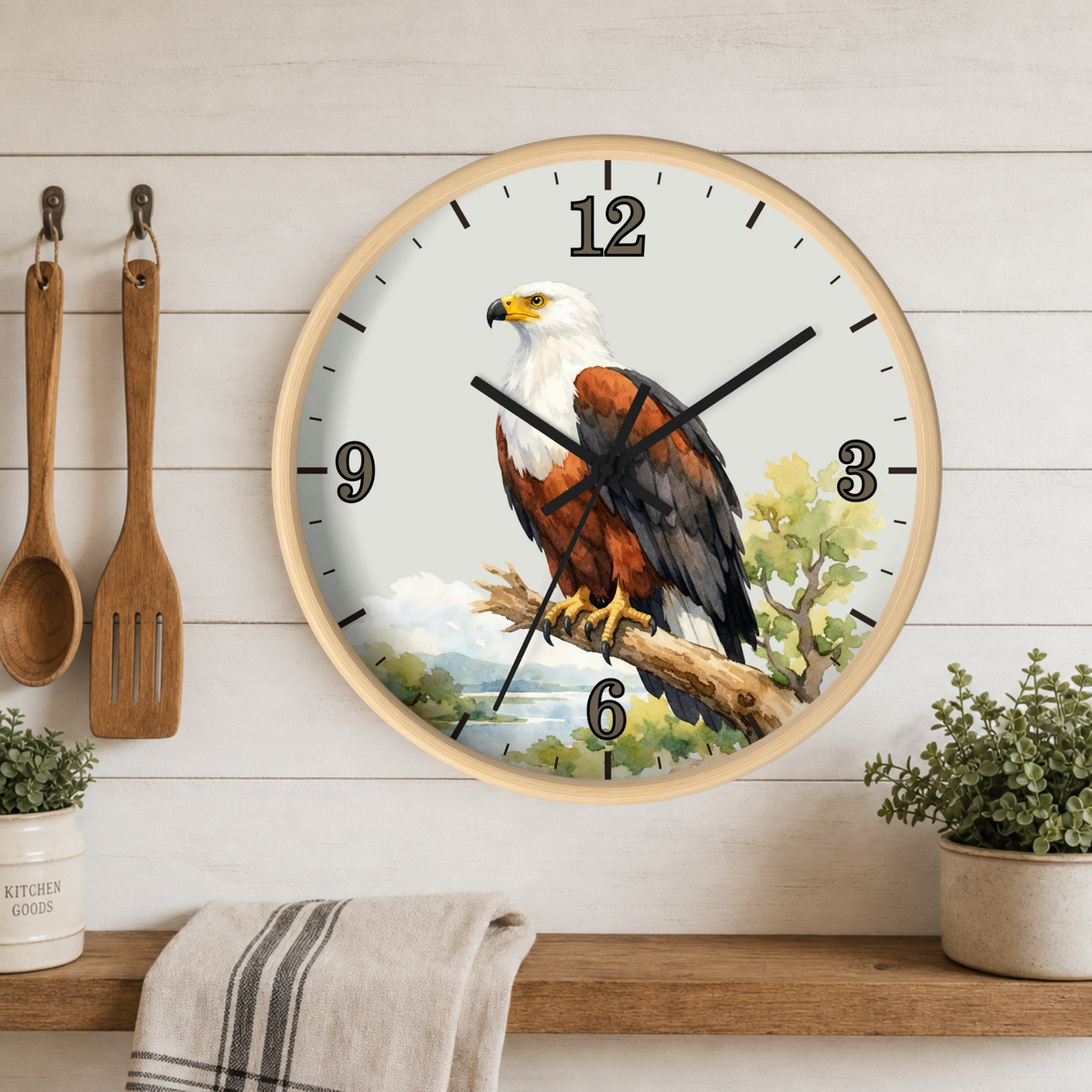 A round wall clock with a light wood frame hangs on a white wood-paneled wall above a wooden shelf. The clock face features a detailed illustration of a fish eagle perched on a branch, with black Arabic numerals and black clock hands.