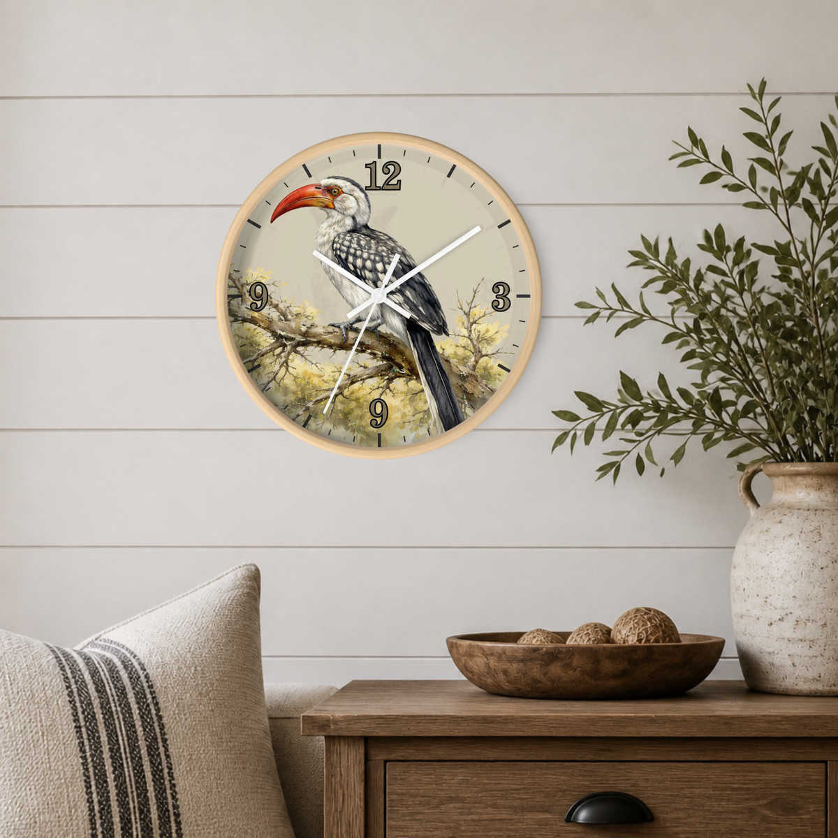 A round wall clock with a natural wood frame hangs on a white shiplap wall above a wooden side table. The clock face features a detailed illustration of a bird perched on a branch, with black Arabic numerals and white hands.