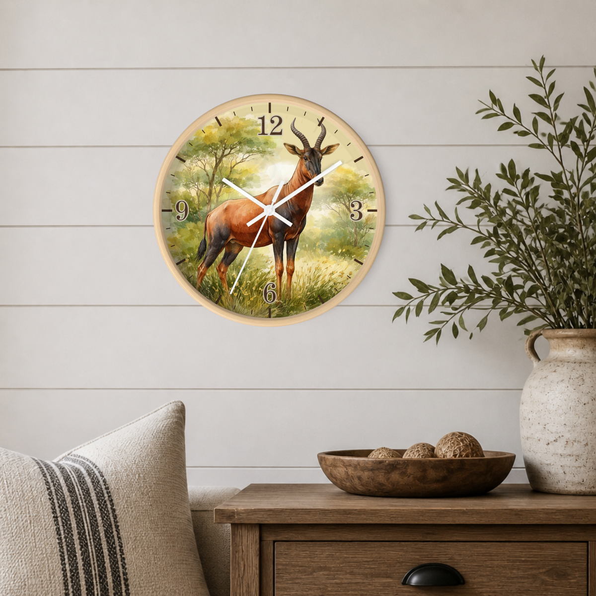 A round wall clock with a natural wood frame hangs on a white shiplap wall above a wooden side table. The clock face features a painted scene of a brown antelope in a grassy landscape with trees, and the numbers are printed in black.