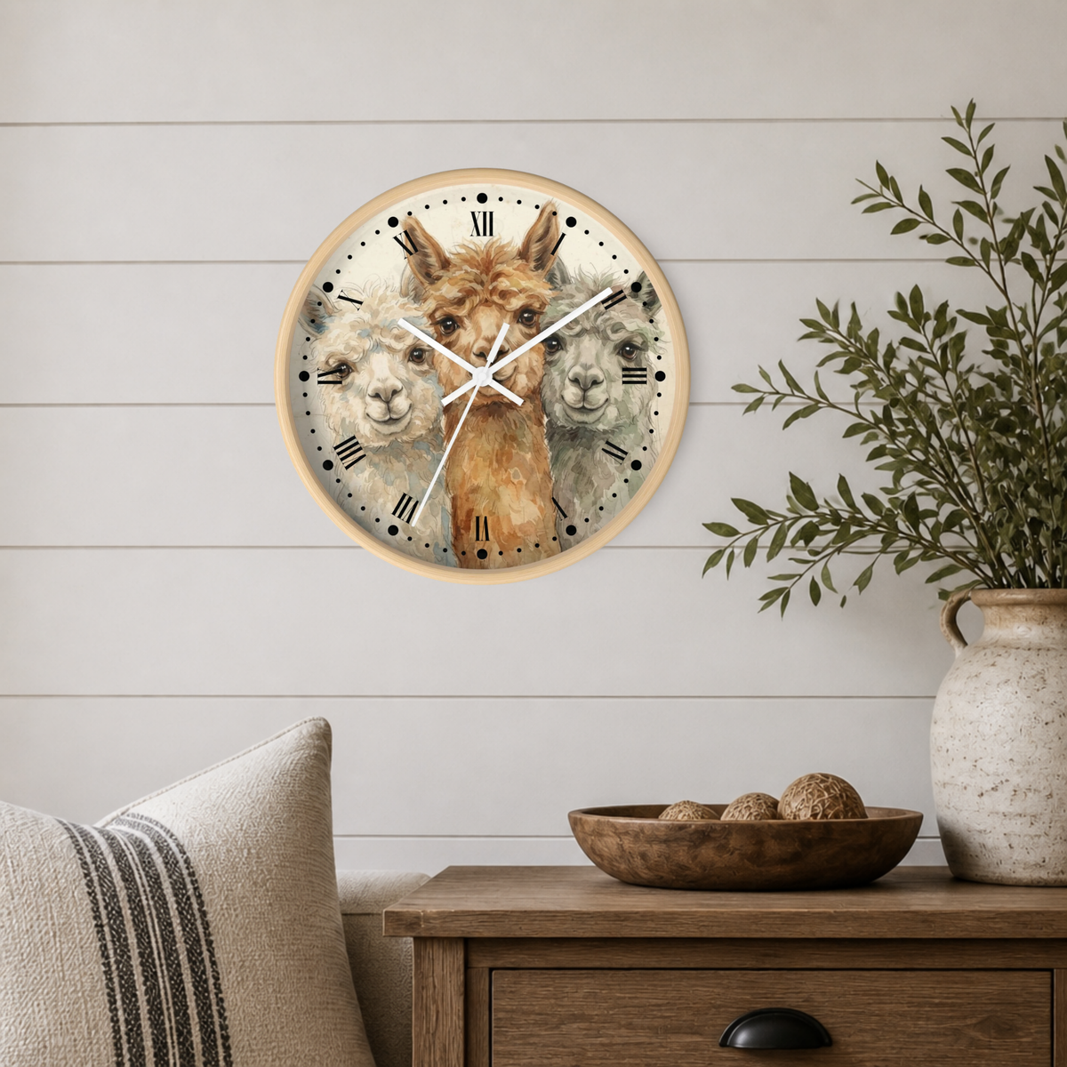 Round wall clock with a light wood frame featuring a printed illustration of three alpacas, styled on a white shiplap wall above a wooden side table with a plant and decorative bowl.