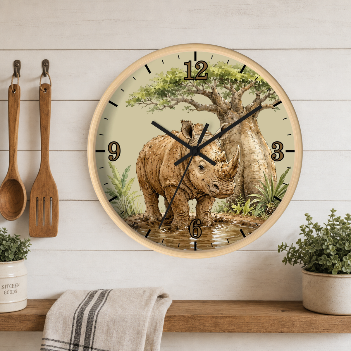 Round wall clock with a light wood frame featuring a detailed illustration of a rhinoceros under a tree on the clock face. The clock is mounted on a white wood-paneled wall above a wooden shelf with kitchen utensils, a towel, and small potted plants.