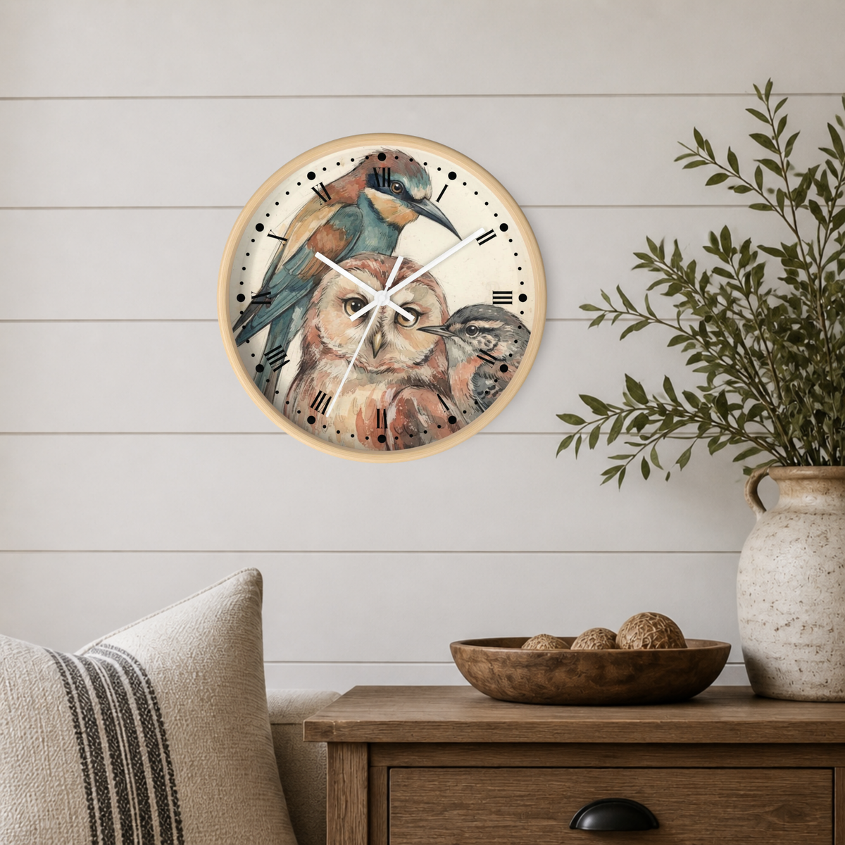 A round wall clock with a watercolor owl and bird design on the face, mounted on a white shiplap wall above a wooden side table with a vase and bowl.