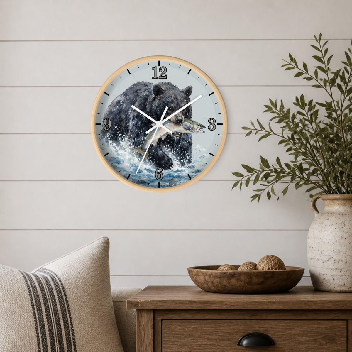 A round wall clock with a light wood frame hangs on a white shiplap wall above a wooden side table. The clock face features a detailed illustration of a black bear in water catching a fish, with black Arabic numerals serving as hour markers.