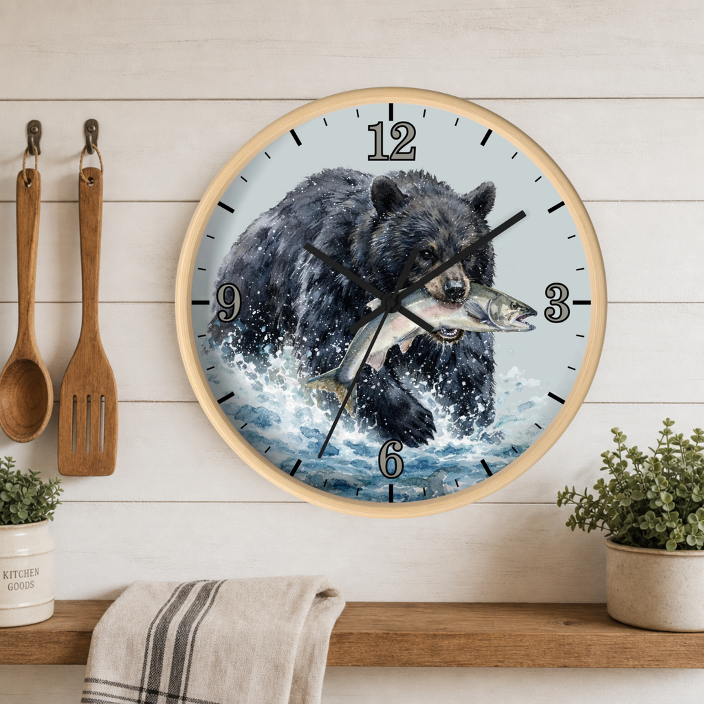 Round wall clock with a light wood frame featuring an illustration of a bear catching a fish, displayed on a white wood plank wall above a shelf with kitchen utensils and plants.