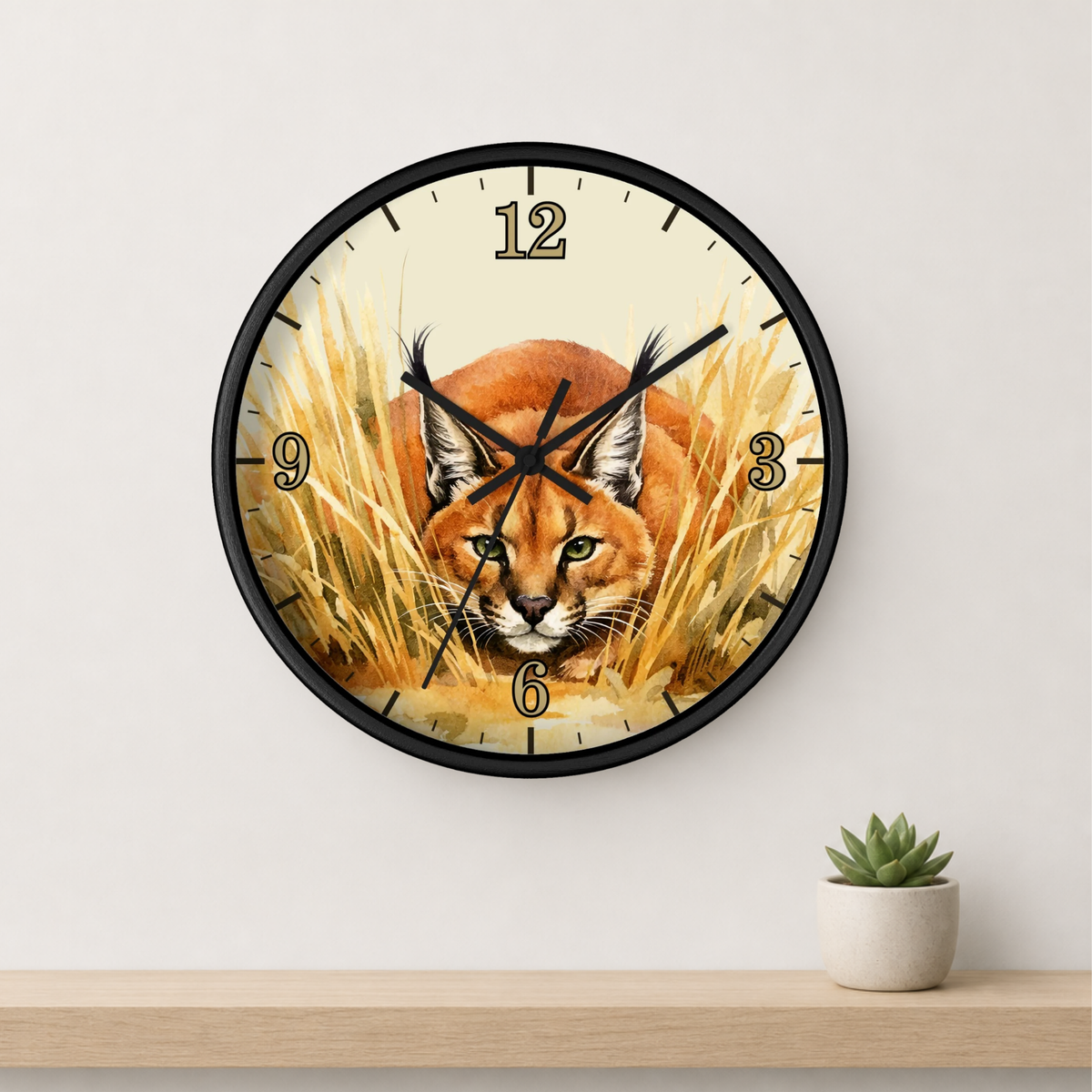 A round wall clock with a black frame and a printed illustration of a caracal in golden grass, mounted on a light gray wall above a wooden shelf with a small potted succulent.