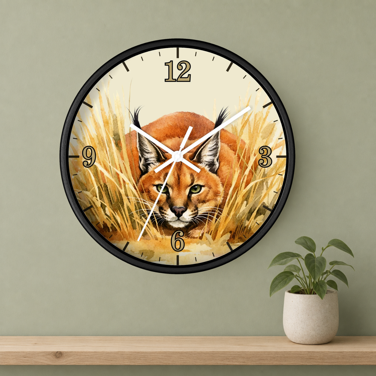 Round wall clock with a black frame and a printed illustration of a lynx cat in golden grass, mounted on a sage green wall above a wooden shelf with a small potted plant.
