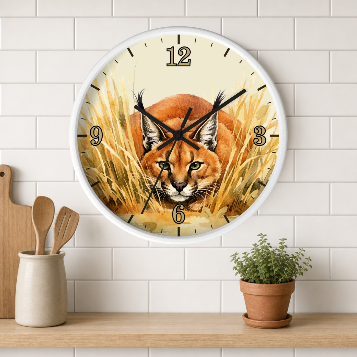 A round wall clock with a white frame hangs on a white subway tile wall above a wooden shelf. The clock face features a detailed illustration of a caracal cat in golden grass.