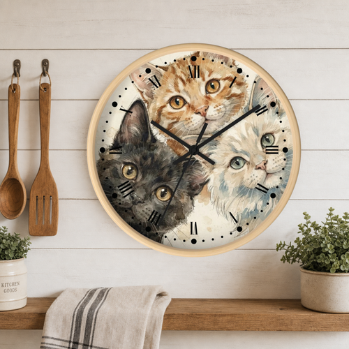 A round wall clock with a light wood frame featuring a watercolor-style illustration of three cats' faces. The clock is mounted on a white wood-paneled wall above a wooden shelf with kitchen utensils, a towel, and potted plants.
