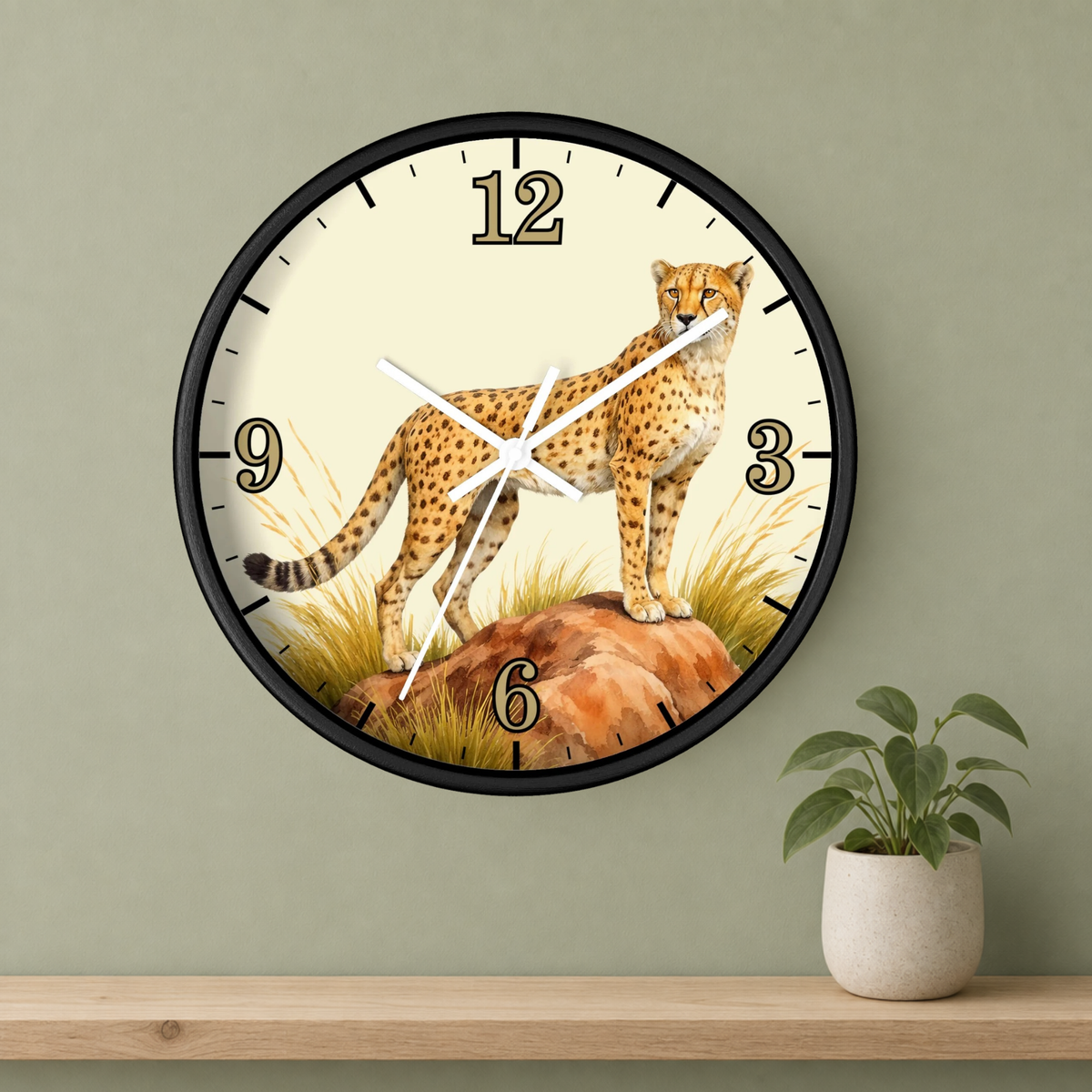 A round wall clock with a black frame and a safari design featuring a cheetah standing on rocks with grass, mounted on a sage green wall above a wooden shelf with a small potted plant.