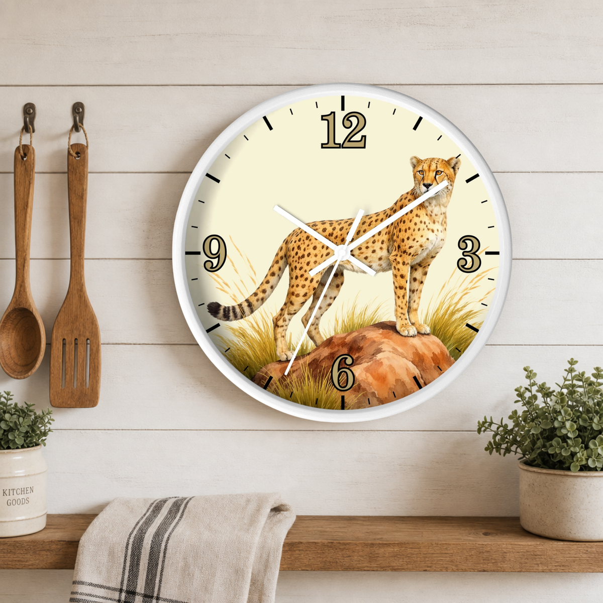 A round wall clock with a white frame and a safari-themed face featuring a cheetah standing on rocks. The clock is mounted on a light wood paneled wall above a wooden shelf with kitchen utensils, a towel, and small potted plants.