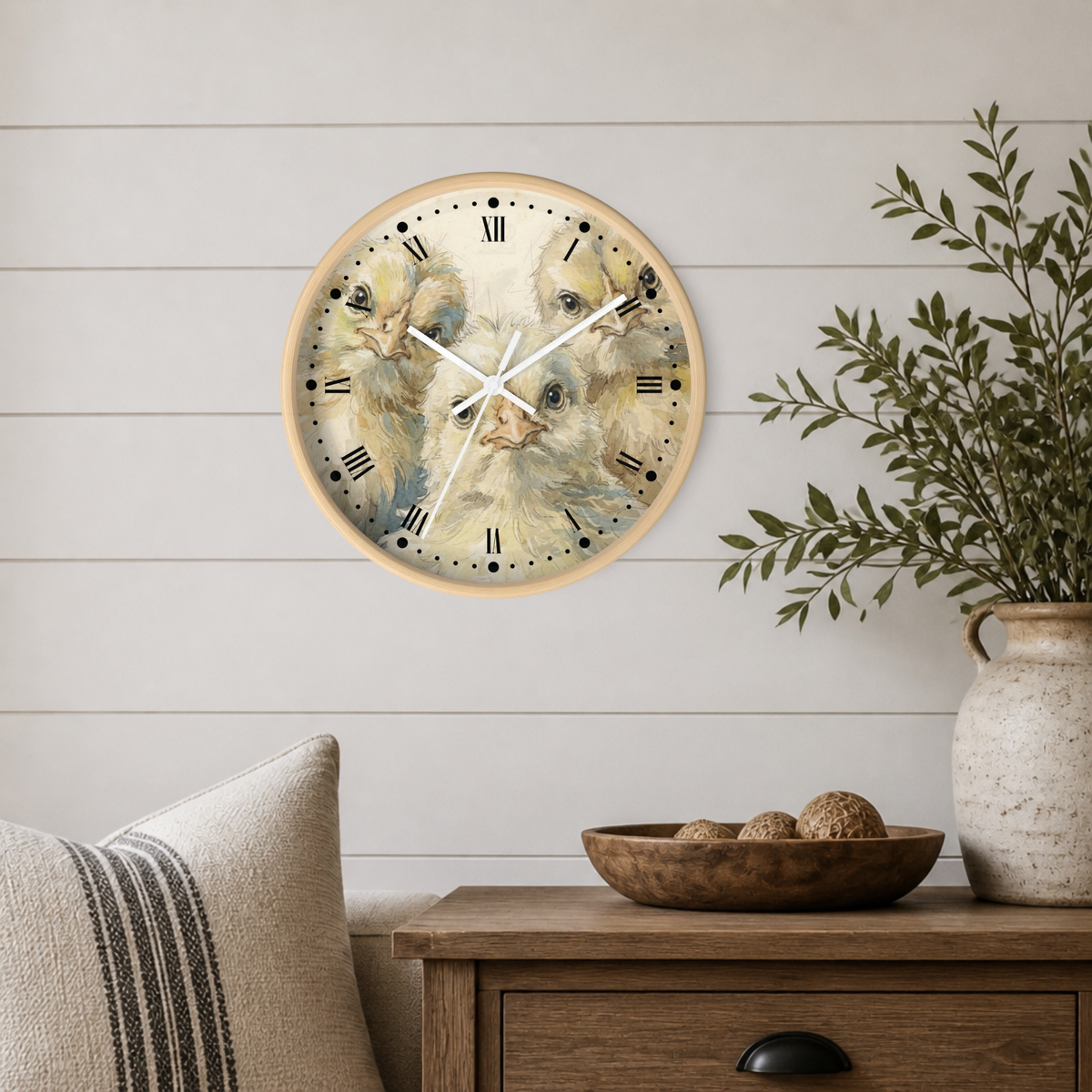 Round wall clock with a light wood frame and a detailed illustration of three yellow chicks on the face, mounted on a white shiplap wall above a wooden side table with a plant and decorative bowl.